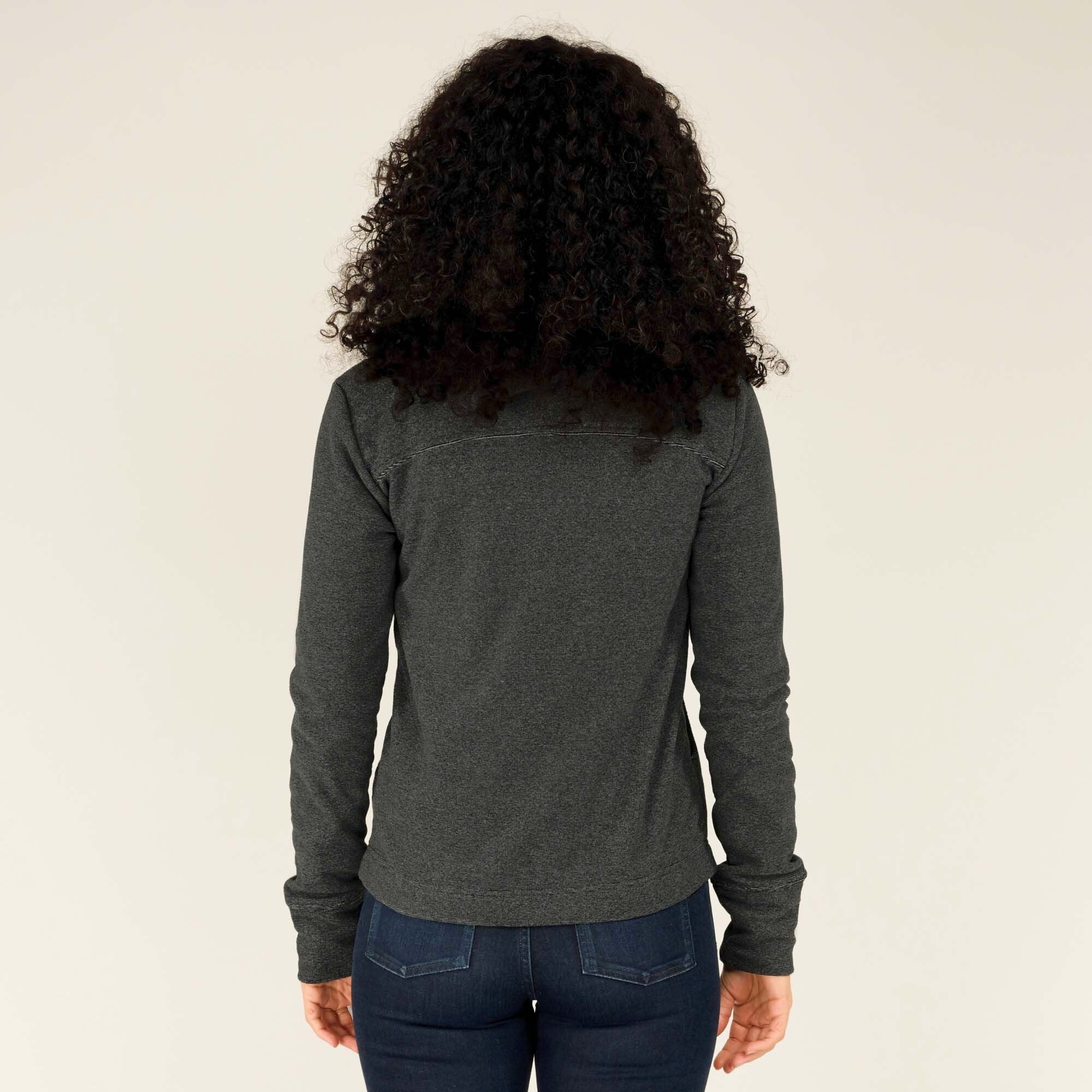 Back view of the model showcasing the rear design of the Sherpa Adventure Gear Rolpa Full Zip Hoodie in Grey. The hoodie has a simple, smooth back panel with the same subtle texture, and the hood rests flat against her shoulders. She continues to wear dark jeans and has her hands at her sides.