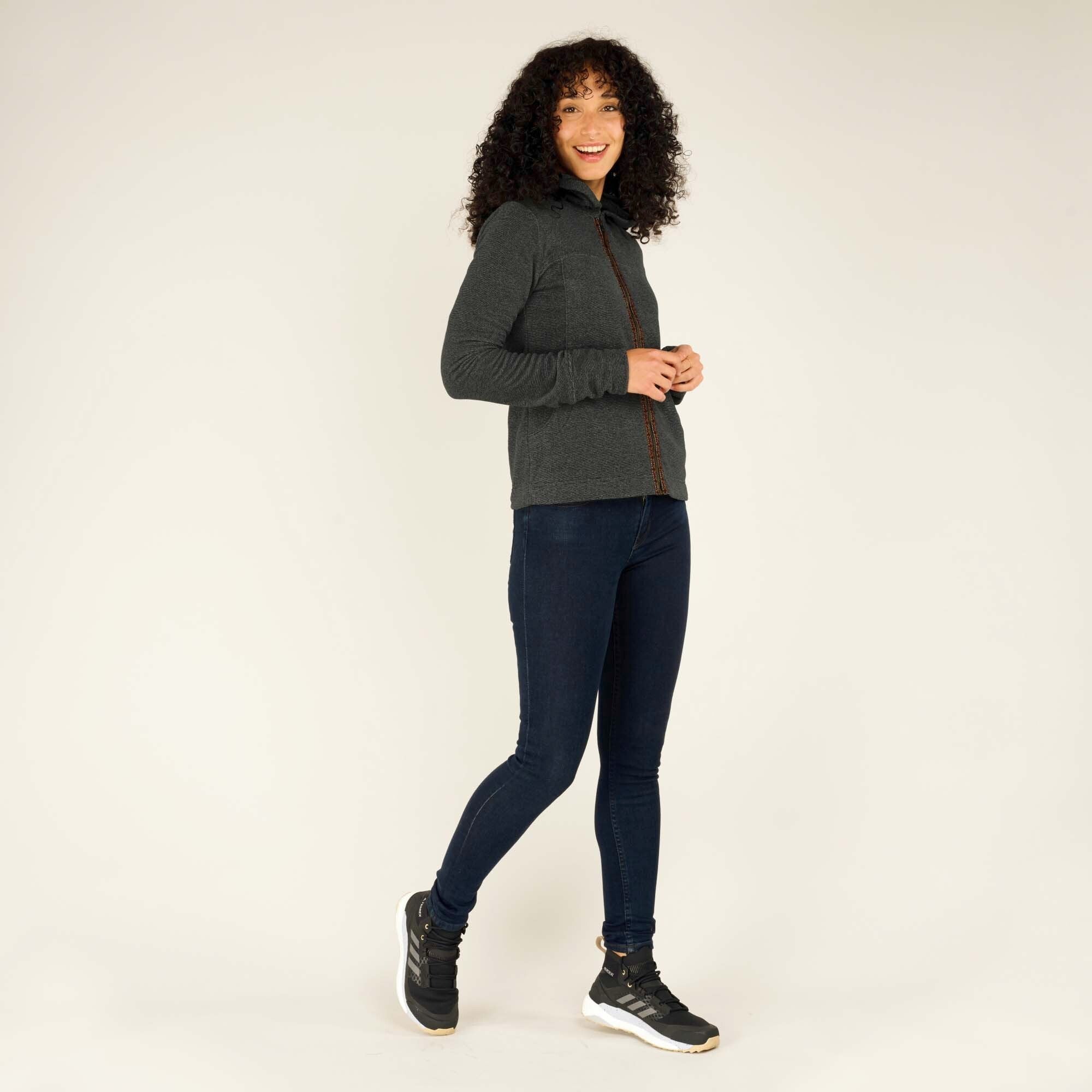 The model is mid-stride, smiling and looking over her shoulder while wearing the Sherpa Adventure Gear Rolpa Full Zip Hoodie in Grey. This image highlights the side profile, the slim fit of the hoodie, and the movement-friendly design. She pairs it with dark blue jeans and black sneakers.