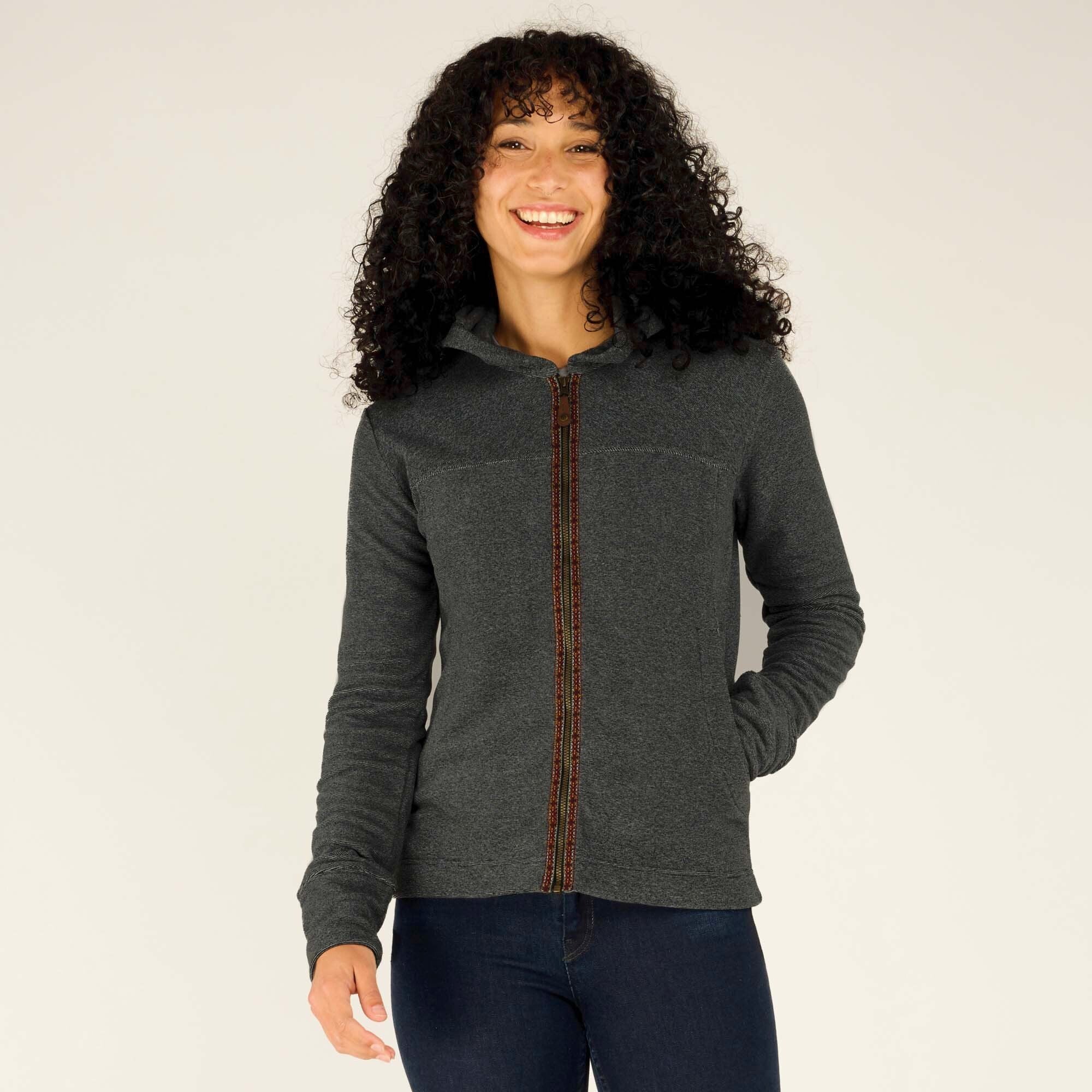 Close-up of the model smiling in the Sherpa Adventure Gear Rolpa Full Zip Hoodie in Grey. The decorative trim along the zipper, the soft texture, and the fitted sleeves are clearly visible. She casually has her hands in the hoodie's pockets.