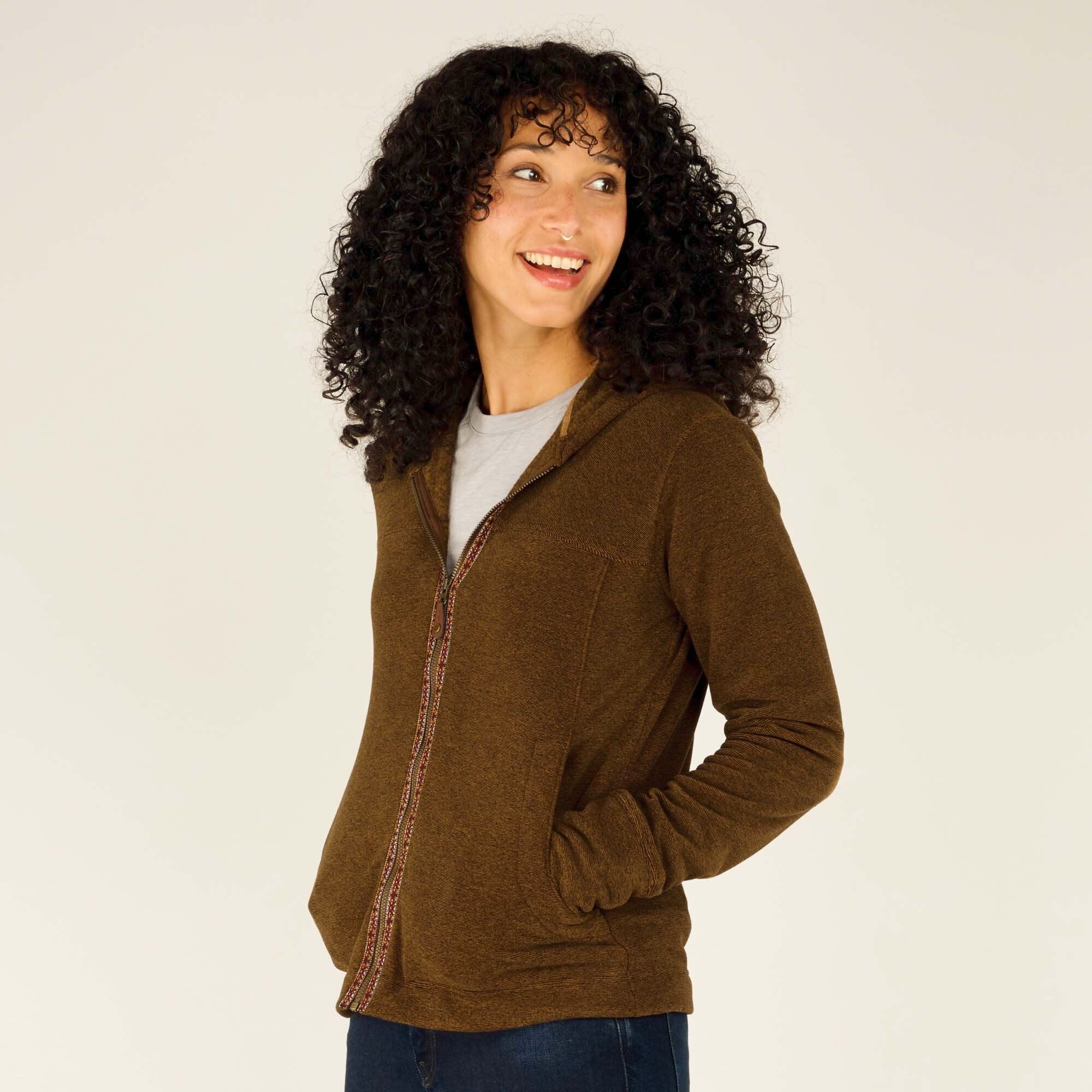 A full-body image of the woman standing in a three-quarter view, showcasing the fit of the Sherpa Adventure Gear Rolpa Full Zip Hoodie in Light Brown. She is wearing fitted dark blue jeans and black sneakers with white soles. She is looking at the camera with a warm smile.