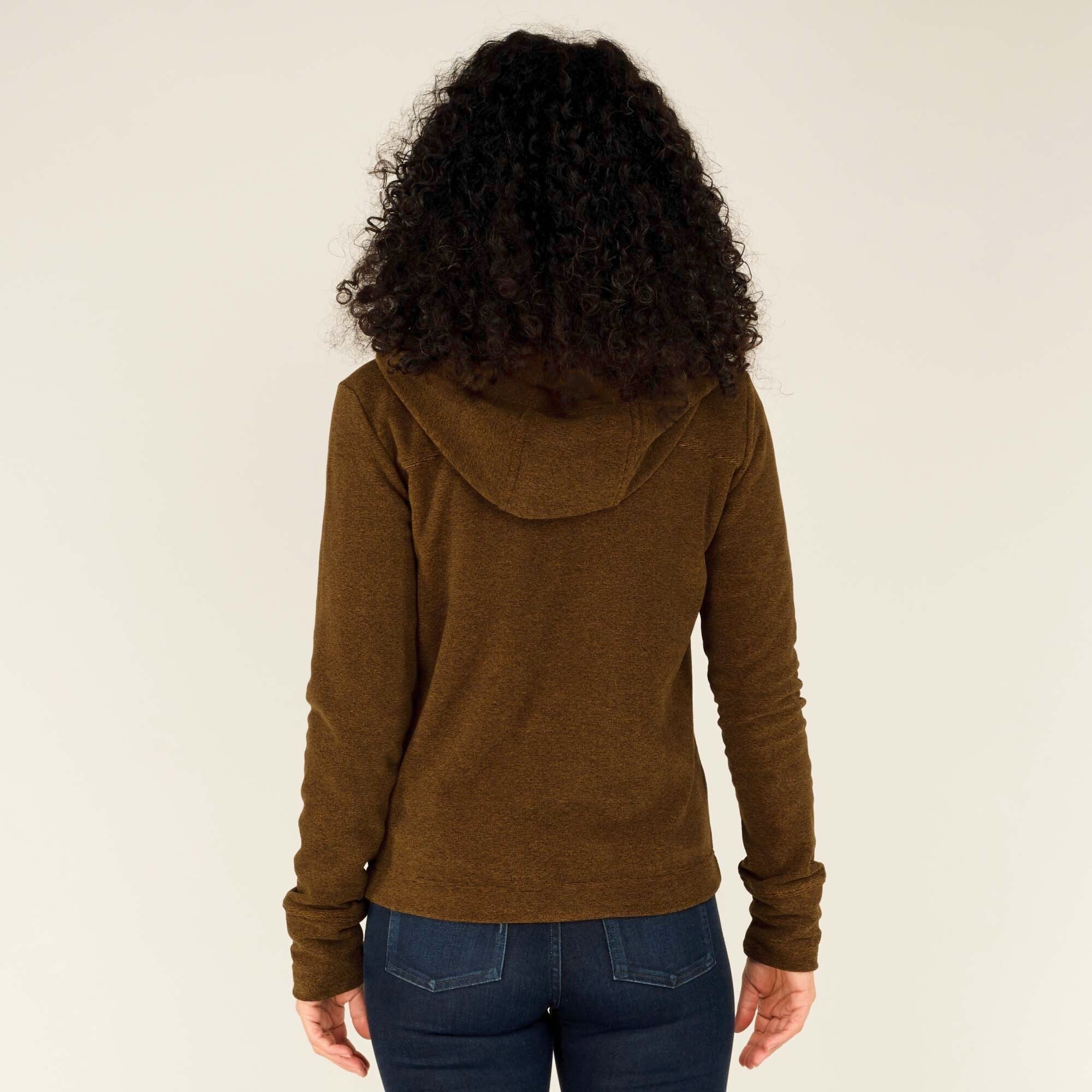 The woman slightly unzips the Sherpa Adventure Gear Rolpa Full Zip Hoodie in Light Brown while holding onto the zipper with one hand. She is looking off to the side with a cheerful expression. The inside of the hood is slightly visible, showing a soft lining. The woven trim along the zipper stands out against the brown fabric.