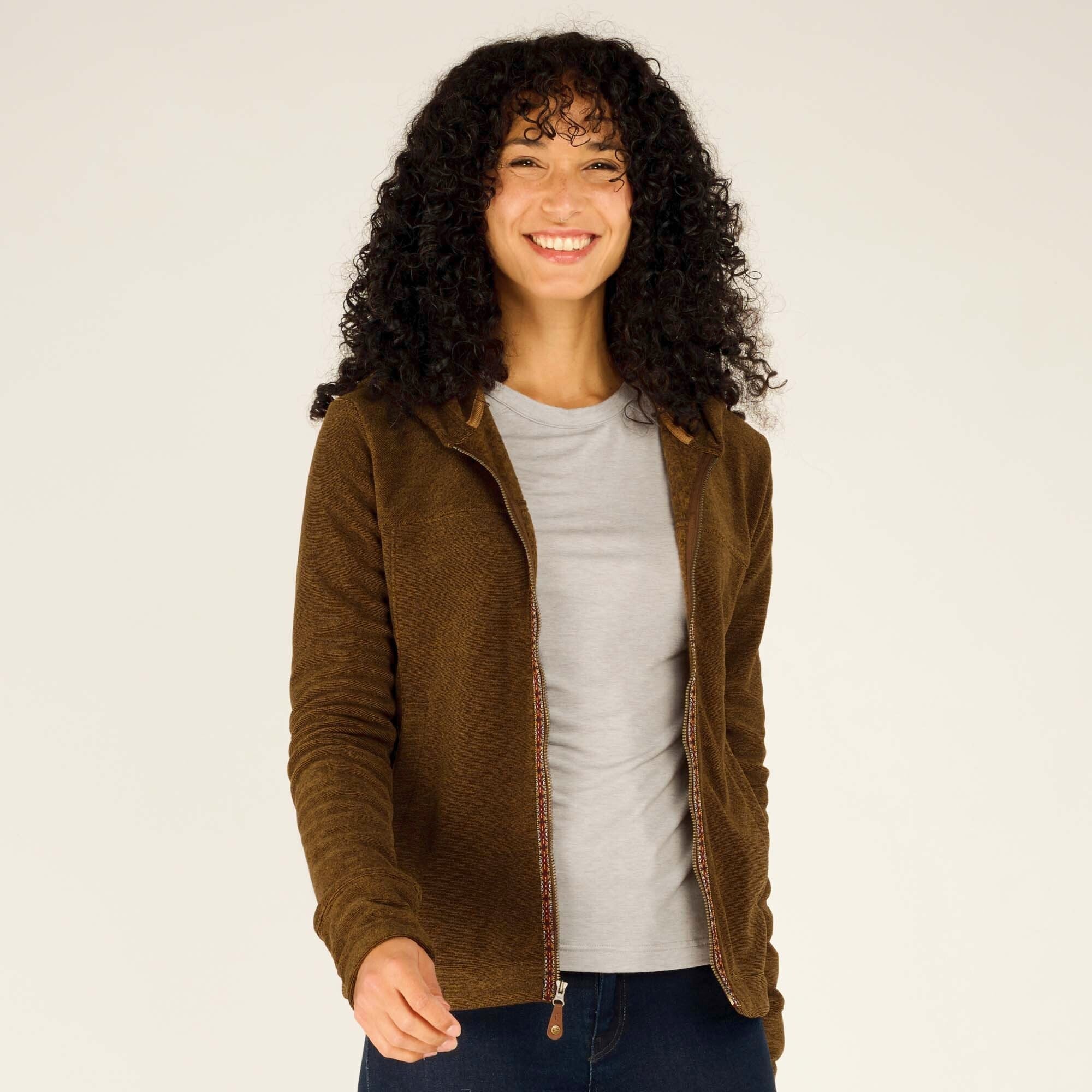 A centered shot of the woman standing with her hands slightly extended in front of her, wearing the zipped-up Sherpa Adventure Gear Rolpa Full Zip Hoodie in Light Brown. She has a relaxed expression and a gentle smile. The fabric texture and trim details are clearly visible.