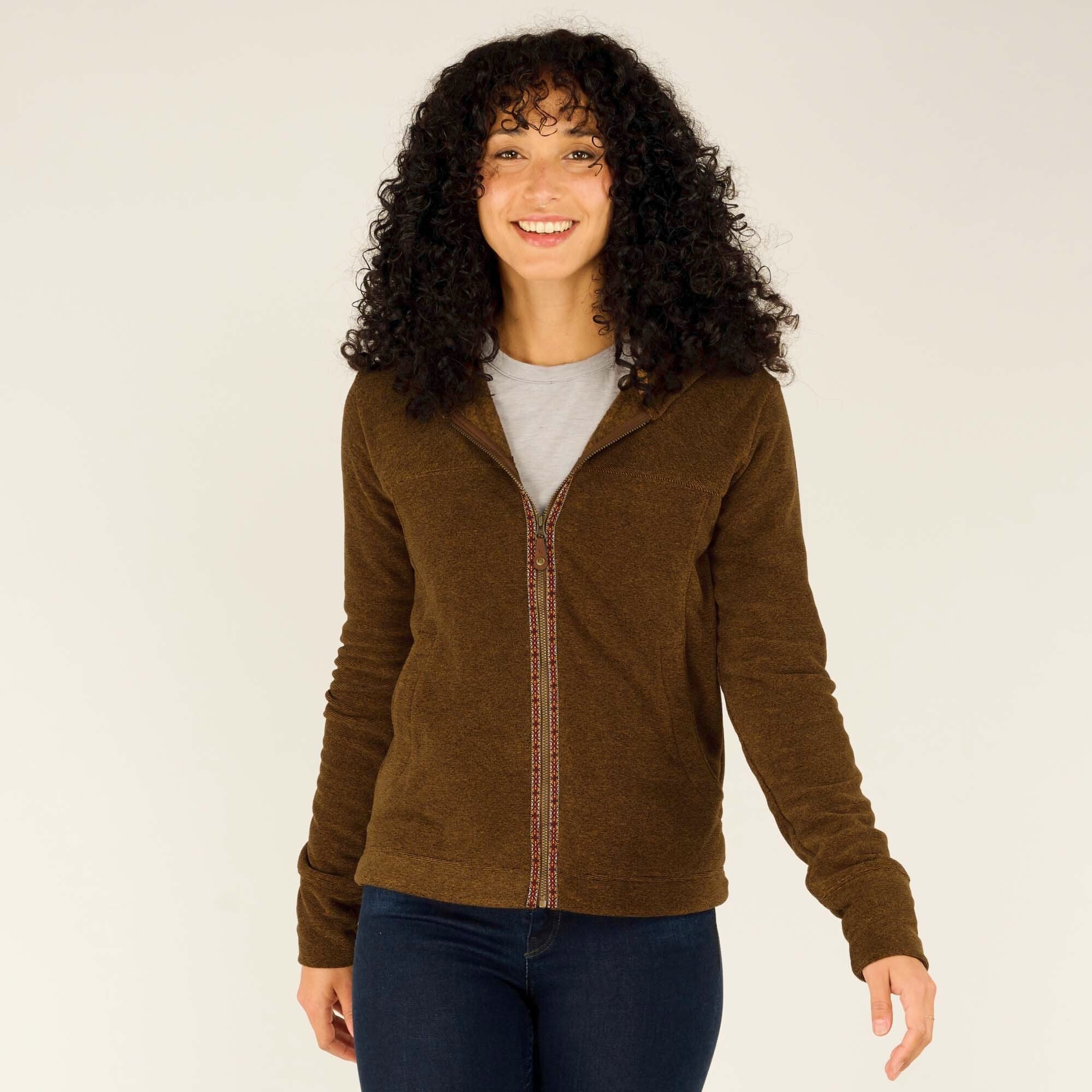 A woman with curly black hair smiles while wearing a Sherpa Adventure Gear Rolpa Full Zip Hoodie in Light Brown with a hood. The hoodie has a textured fabric and features a decorative woven trim along the zipper. She has her hands in the hoodie pockets and is looking off to the side. She is also wearing a gray t-shirt underneath and dark blue jeans. The background is a neutral beige color.