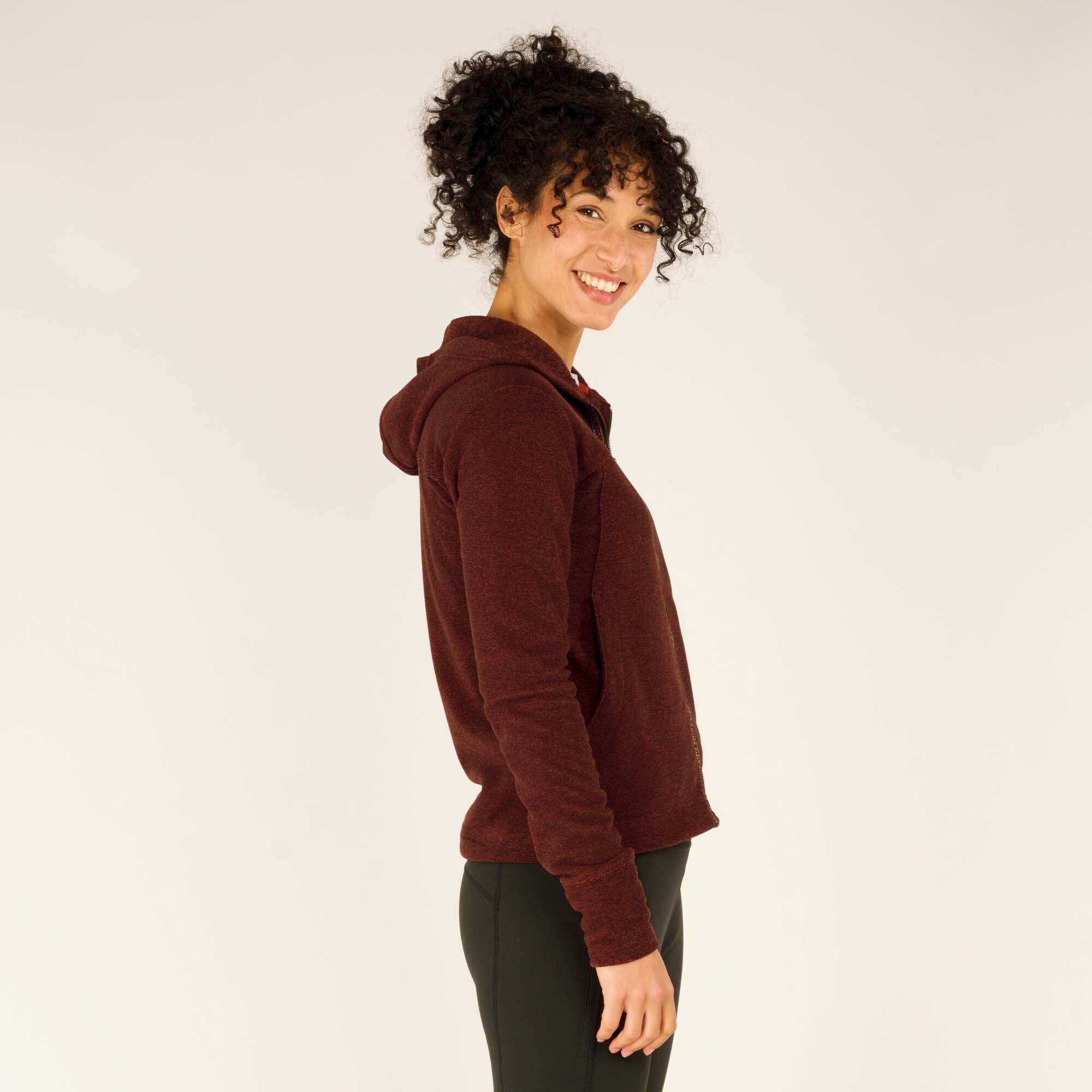 The model wearing the Sherpa Adventure Gear Rolpa Full Zip Hoodie in Red is standing in a relaxed stance with the zipper slightly open, revealing a layer underneath. The sleeves extend to the model’s wrists, and the hem sits just below the waist. The hoodie’s overall structure and casual style are emphasized, making it ideal for layering.