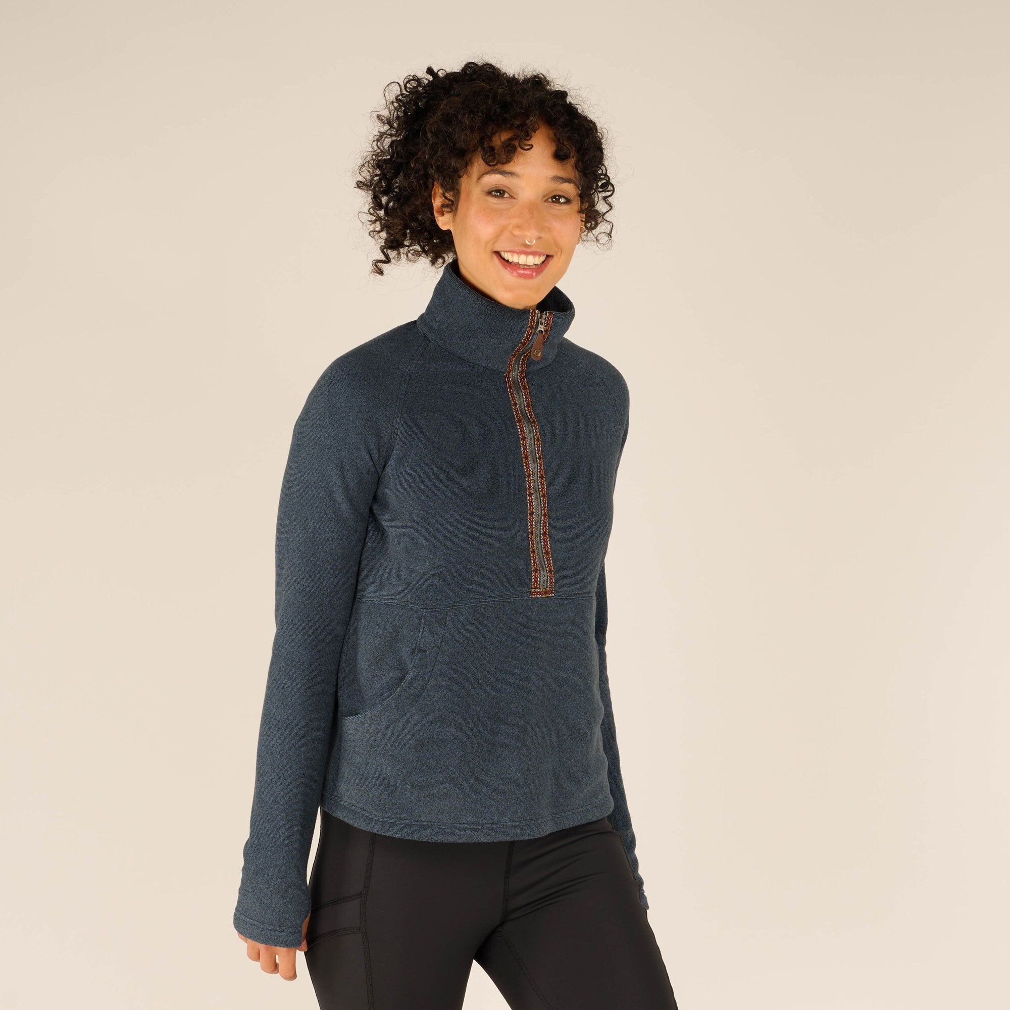 Close-up front view of the woman walking toward the camera, still wearing the Sherpa Adventure Gear Rolpa Half Zip Pullover in Blue. She is smiling brightly and the decorative woven trim along the zipper is clearly visible, adding contrast to the solid blue fleece fabric.