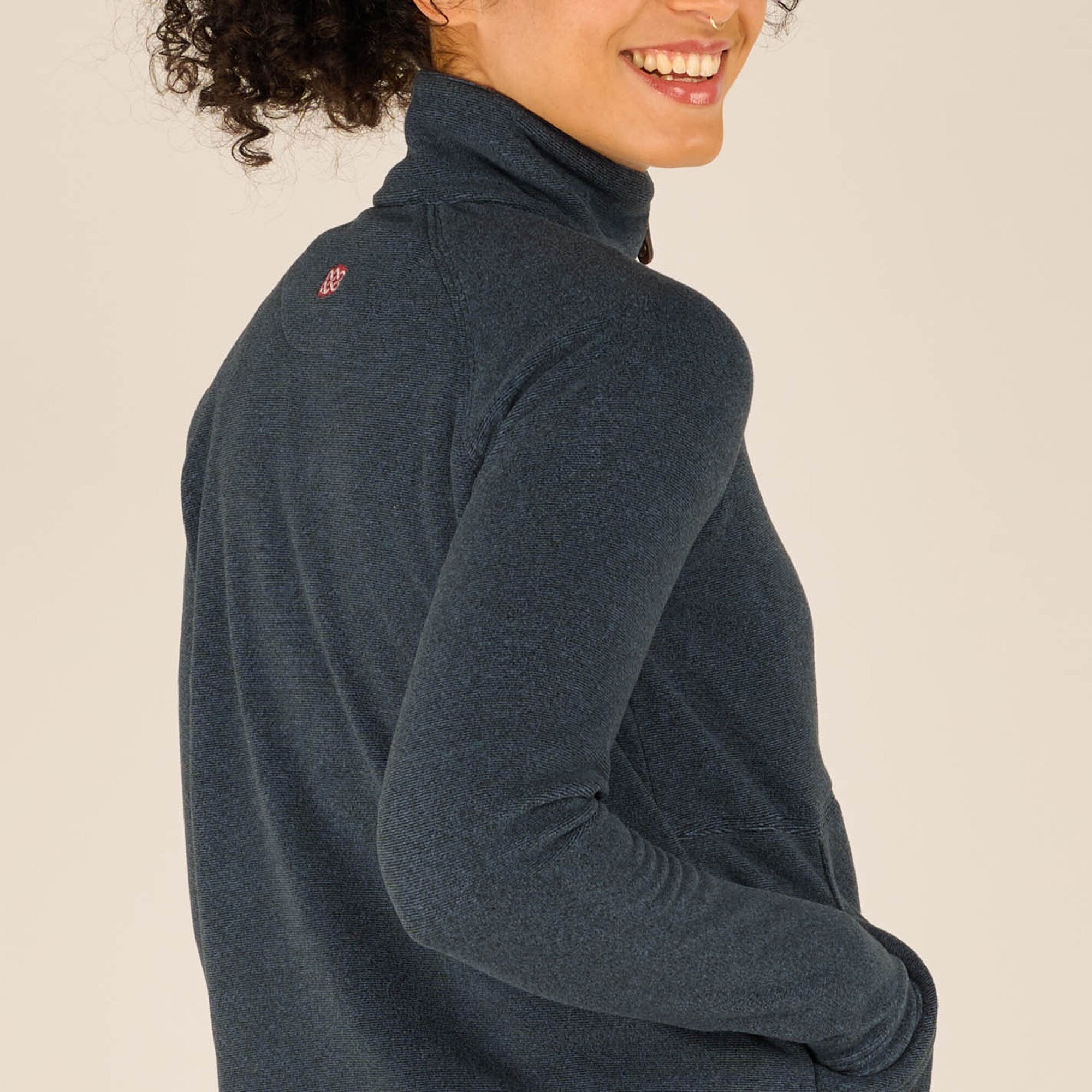 Side angle close-up of the woman turned slightly, smiling over her shoulder. This image highlights the textured fleece fabric of the Sherpa Adventure Gear Rolpa Half Zip Pullover in Blue, the fit of the sleeves, and the subtle embroidered logo at the upper back.