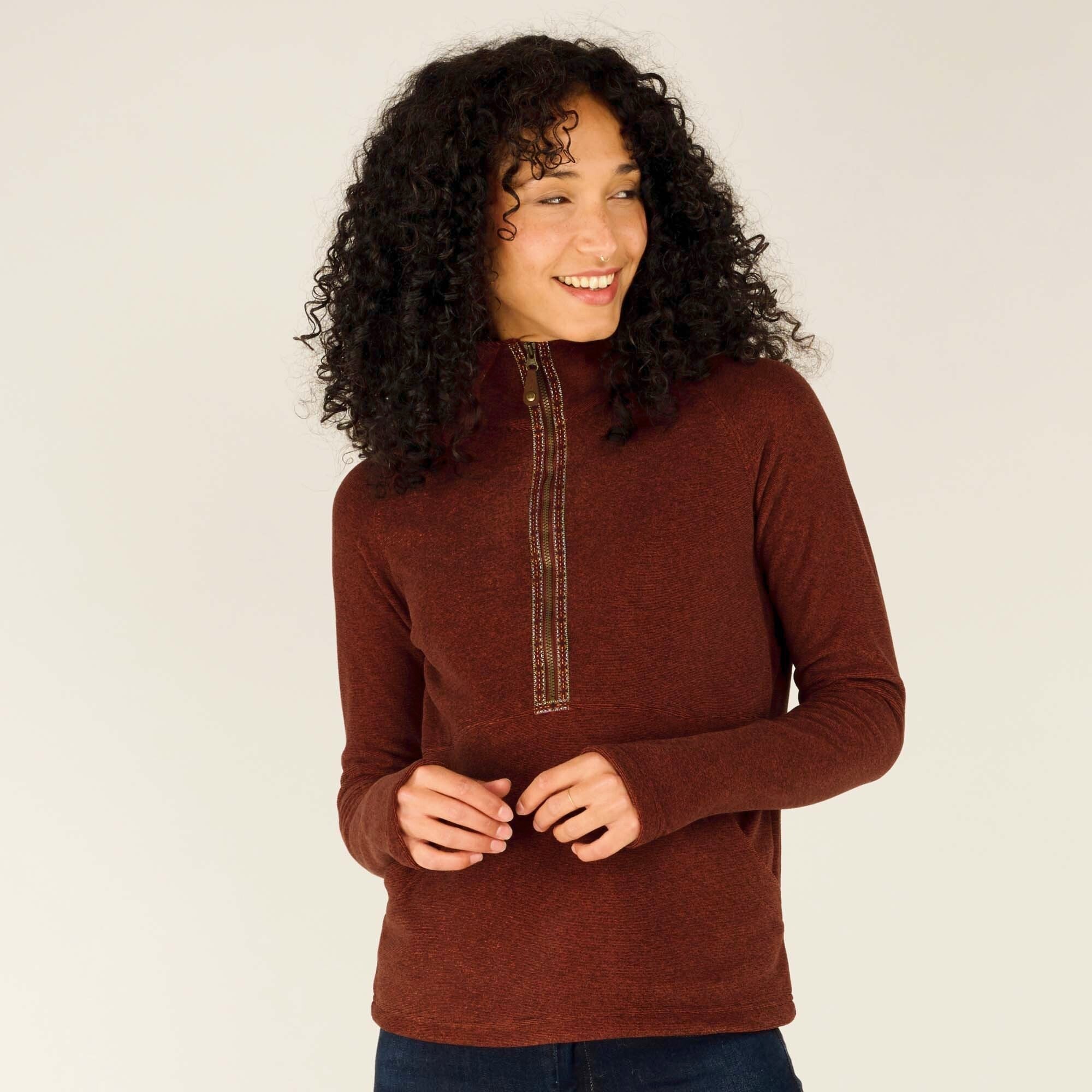 A woman with curly dark hair and medium skin tone is standing facing the camera, wearing a Sherpa Adventure Gear Rolpa Half Zip Pullover in Red. The pullover features a half-zip with a decorative, multi-colored patterned trim running along the zipper. The woman is smiling softly, and her hands are gently clasped together in front of her. The fabric of the pullover appears soft, and the high collar adds warmth and style.