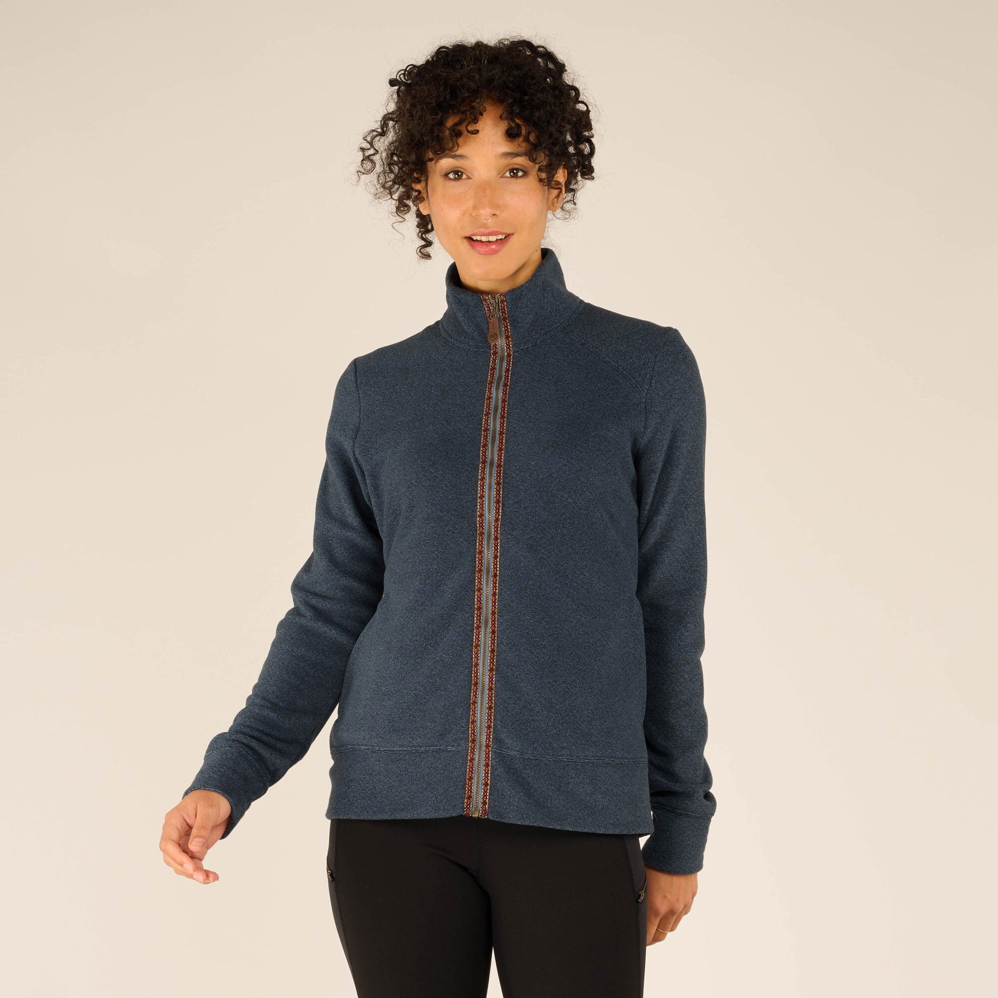 The model stands facing the camera, wearing a Sherpa Adventure Gear Rolpa Jacket in Blue with a high collar. The jacket features a patterned trim along the zipper, adding a decorative detail. The model pairs the jacket with black leggings and black athletic shoes. The neutral background highlights the garment's texture and color.