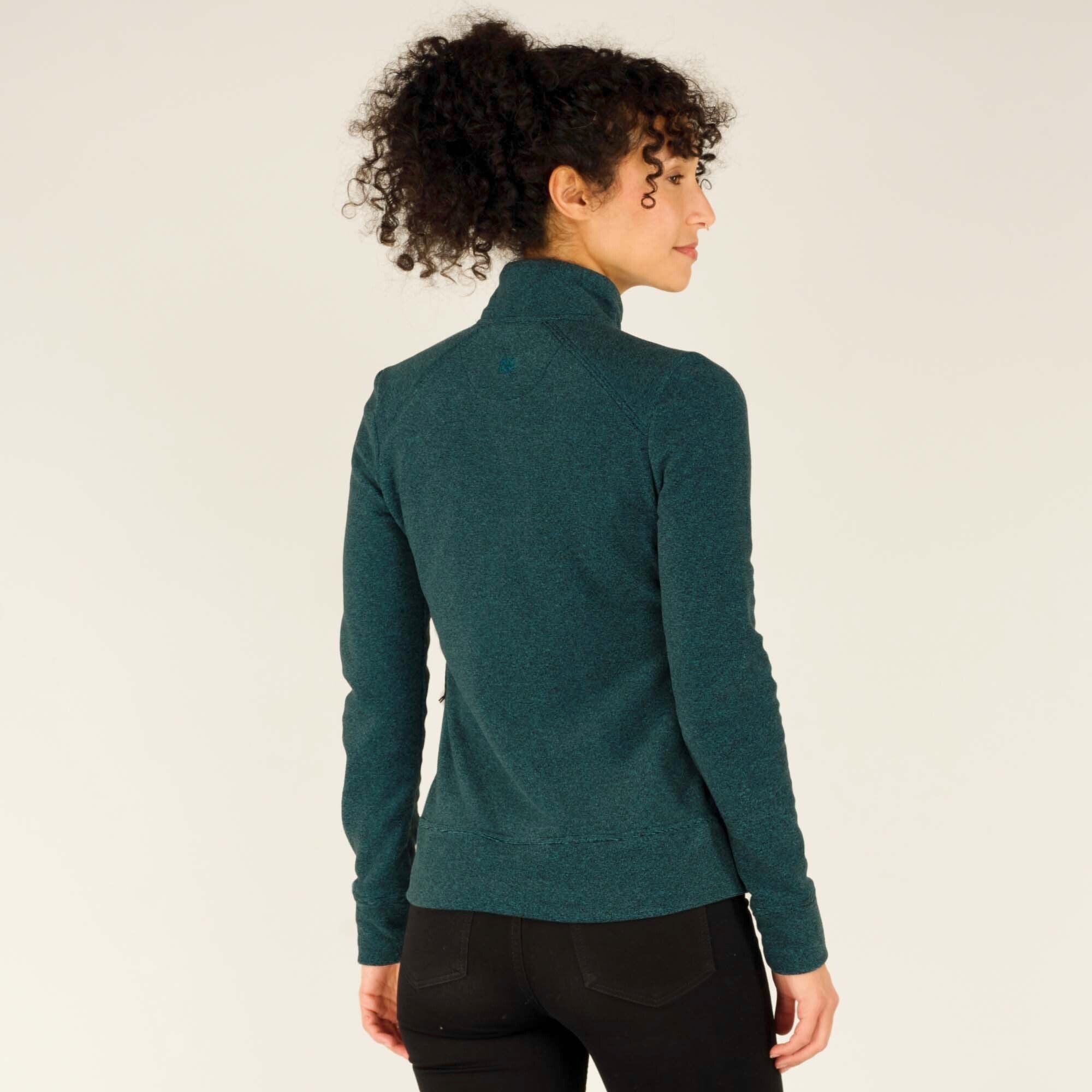 A back view of the woman wearing the Sherpa Adventure Gear Rolpa Jacket in Blue, showing its fit and seam detailing. The high collar is folded slightly, and the fabric has a subtle texture. Her curly hair is pulled up into a loose ponytail, revealing the back of the jacket.