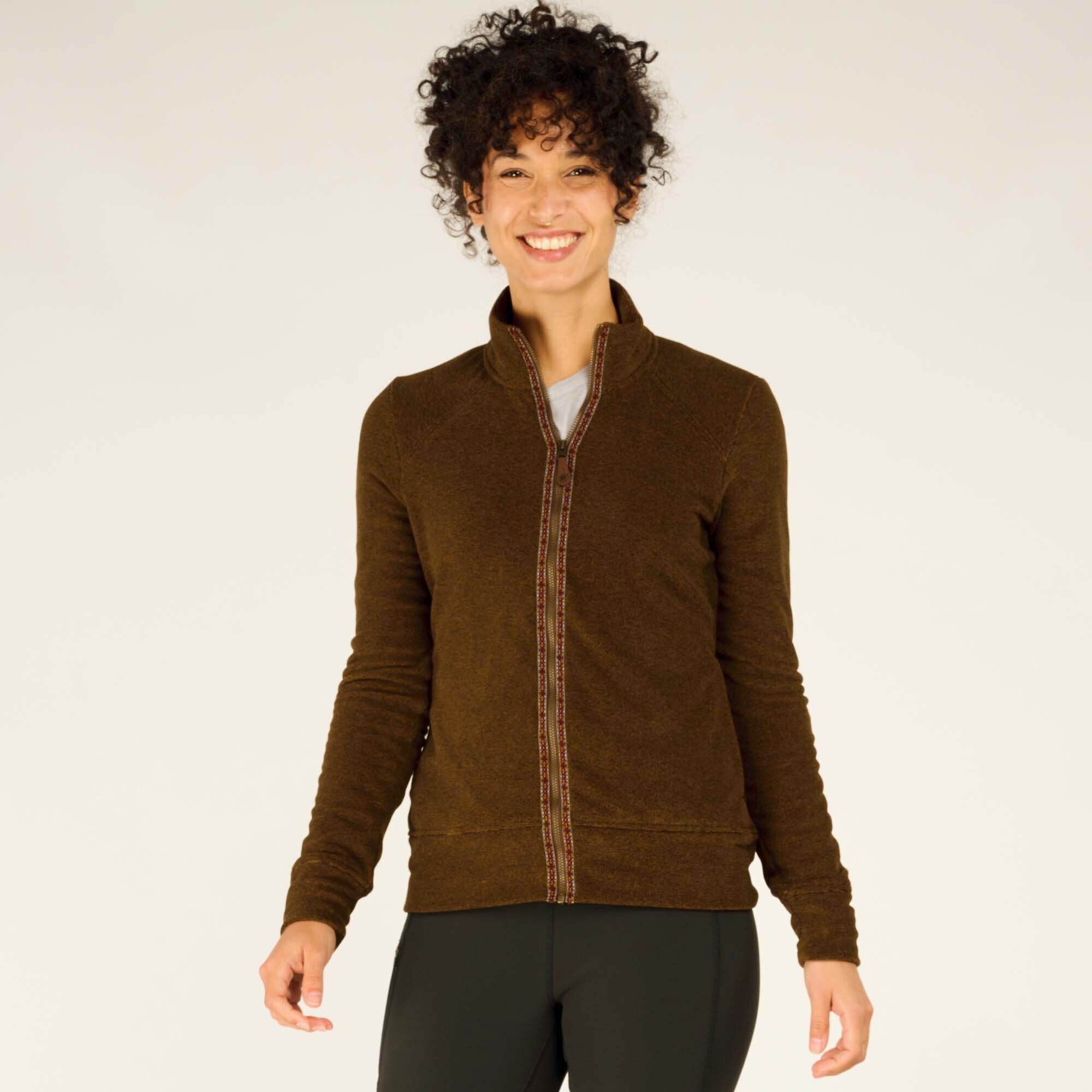 A front view of a woman smiling and wearing a Sherpa Adventure Gear Rolpa Jacket in Light Brown. The jacket features a full-zip front, decorative pattern along the zipper, and a slightly tailored fit. She pairs it with black leggings.