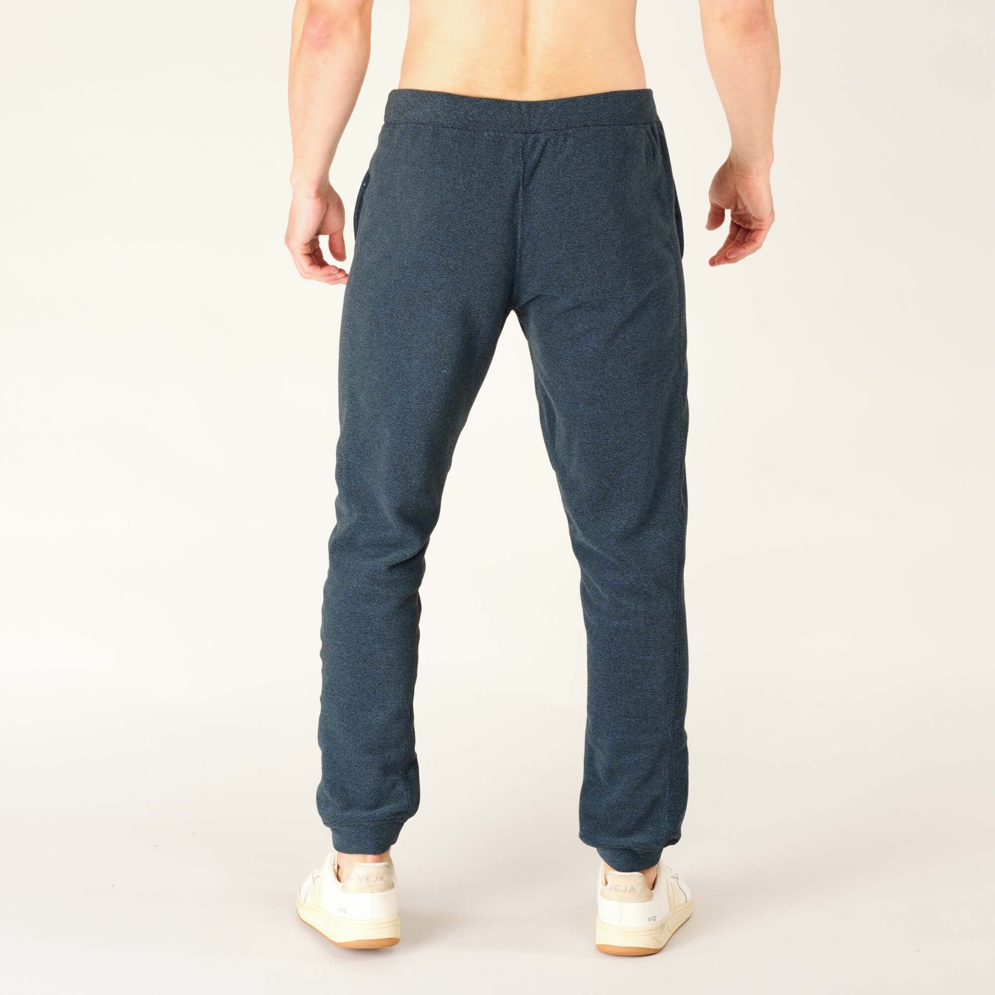 Rear view of the male model wearing the Sherpa Adventure Gear Rolpa Jogger in Blue. The waistband fits comfortably, and the fabric drapes smoothly down the legs. The joggers have two side pockets and simple back detailing. The ankle cuffs are elastic.