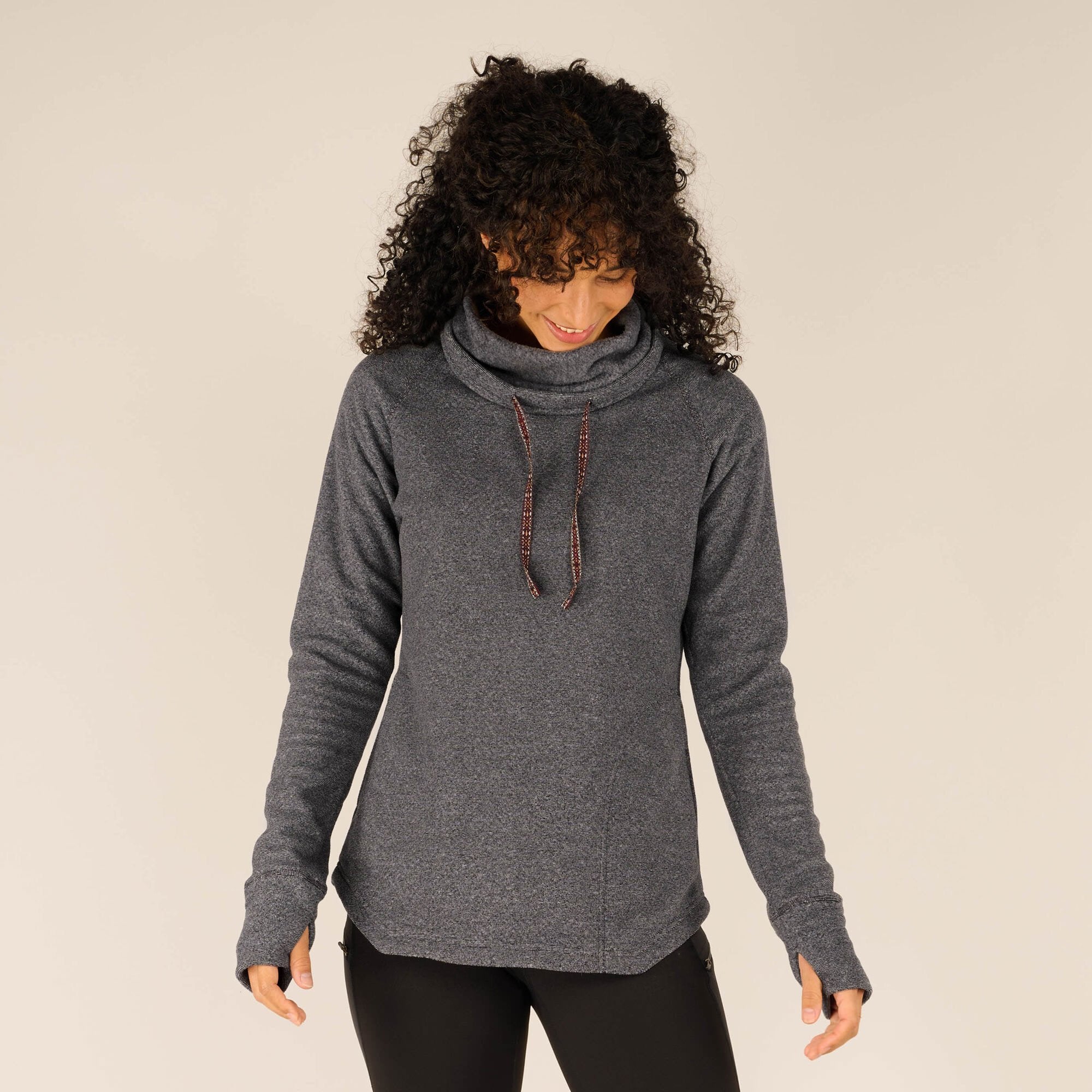 A woman with curly black hair is wearing a Sherpa Adventure Gear Rolpa Pullover in Grey. The pullover features a soft fleece material, a cowl neck with drawstrings, long sleeves with thumb holes, and a slightly curved hemline. She is looking down and smiling slightly while her hands rest at her sides.