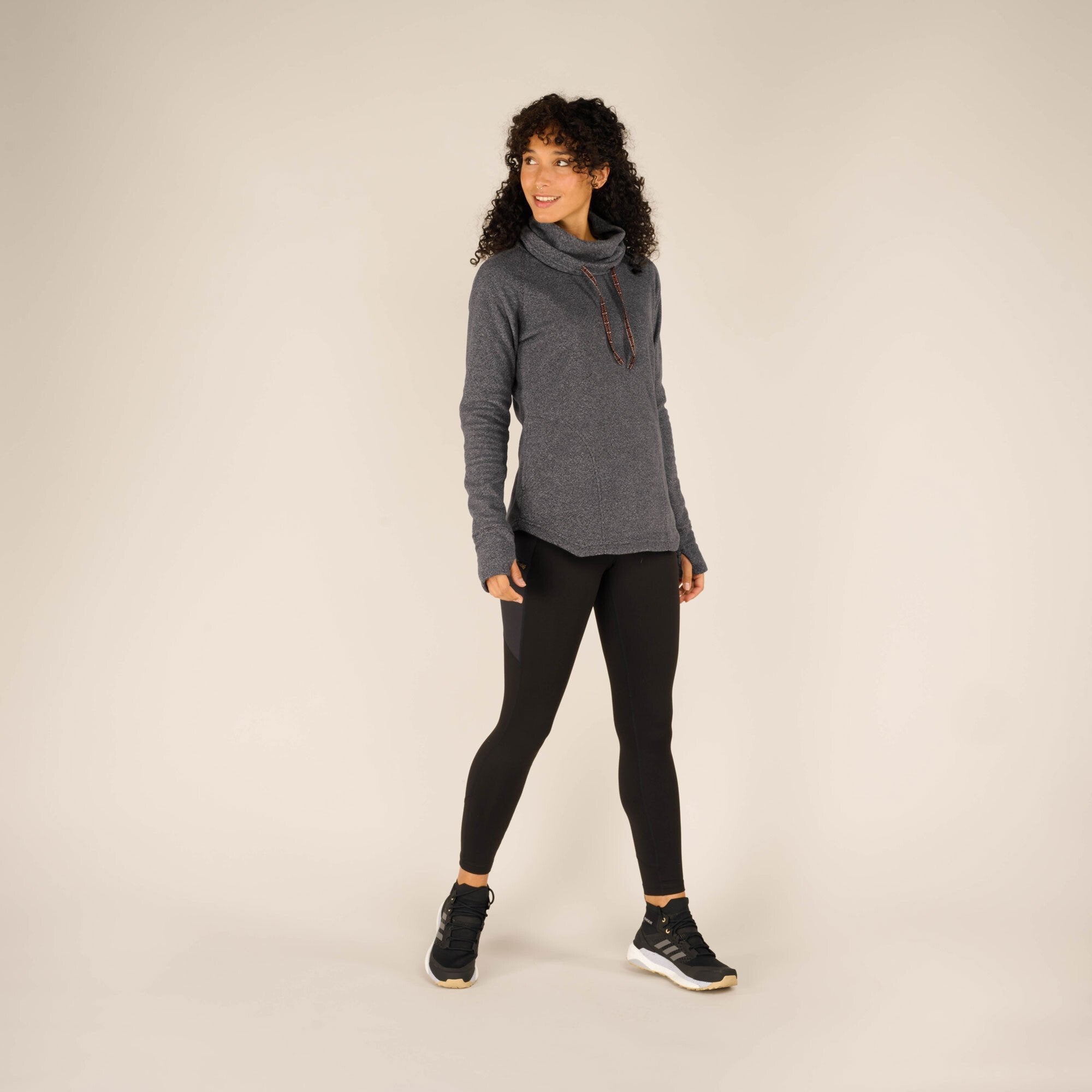 A full-body view of the same woman wearing the Sherpa Adventure Gear Rolpa Pullover in Grey, paired with black leggings and black trainers with white soles. She stands with one foot slightly in front of the other, looking to her right with a relaxed posture against a neutral beige background.