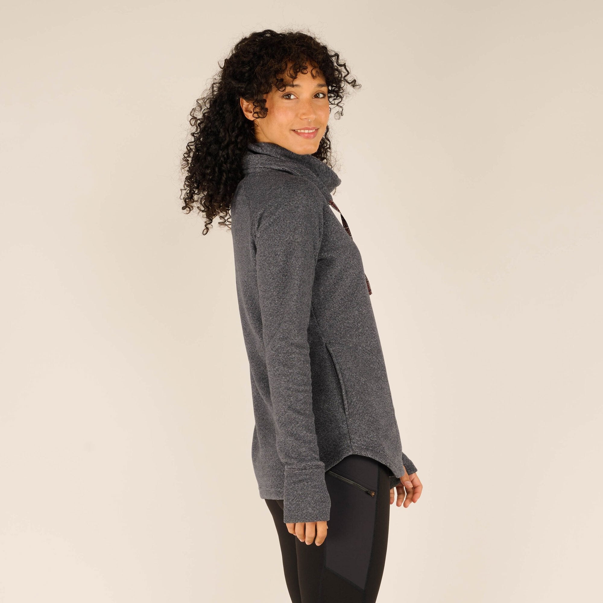 A side profile view of the woman wearing the Sherpa Adventure Gear Rolpa Pullover in Grey, standing with her left shoulder facing the camera. The pullover has a slightly loose fit, and she is smiling gently while glancing over her shoulder.