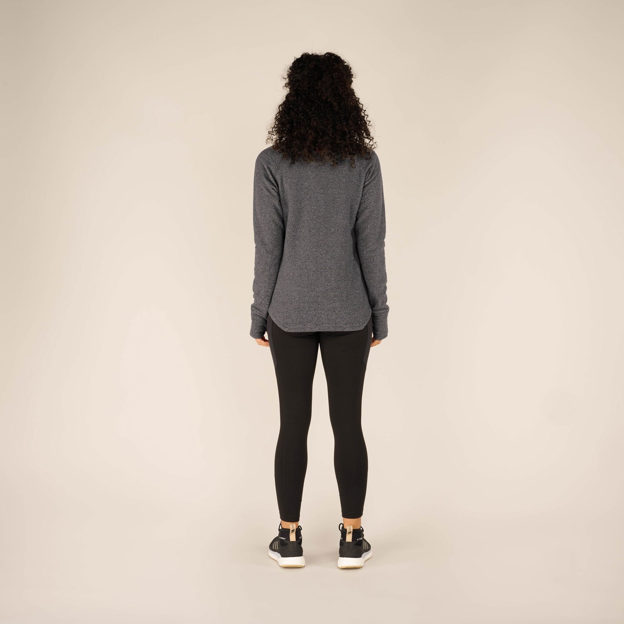 A back view of the woman wearing the Sherpa Adventure Gear Rolpa Pullover in Grey. Her long curly hair cascades down her back, partially covering the cowl neck. The slightly curved hemline and overall relaxed fit of the pullover are visible.