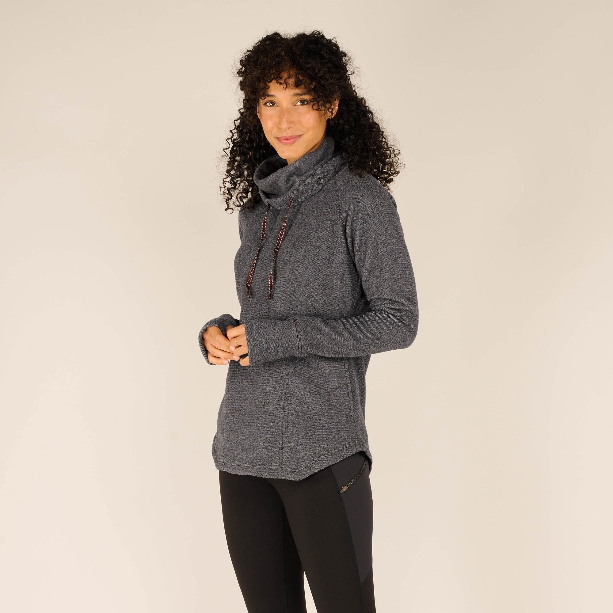 A three-quarter view of the woman slightly turned towards the camera. She is smiling warmly while adjusting the drawstrings of the cowl neck with her hands on the Sherpa Adventure Gear Rolpa Pullover in Grey.