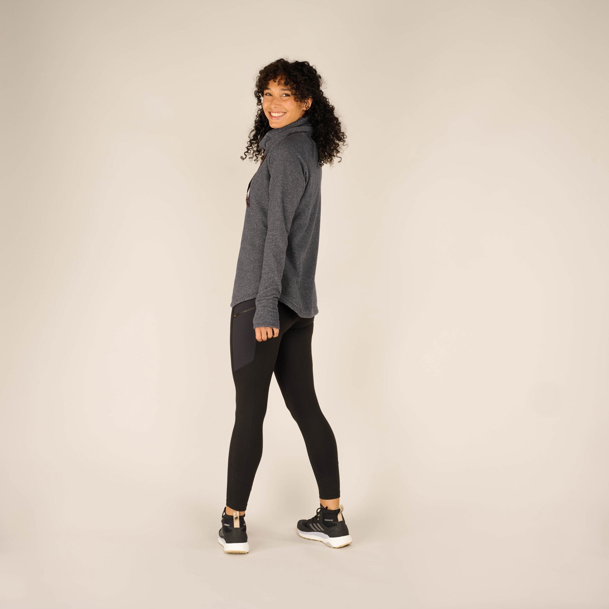 A dynamic side-angle shot of the woman walking, wearing the Sherpa Adventure Gear Rolpa Pullover in Grey. She is smiling brightly while stepping forward, giving a sense of movement. The pullover maintains its relaxed fit, and the thumb holes in the sleeves are visible.