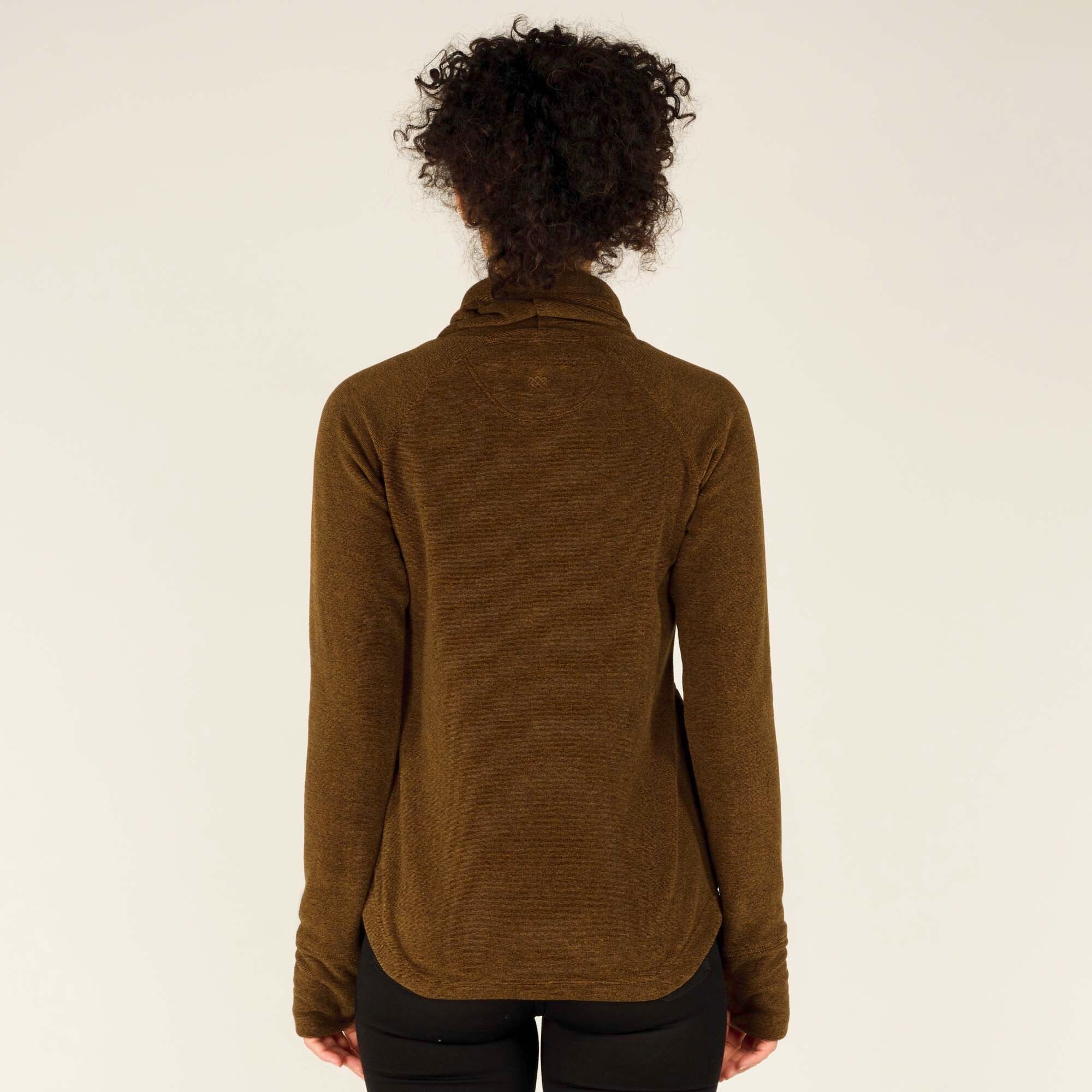 A rear view of the woman showcasing the back of the Sherpa Adventure Gear Rolpa Pullover in Light Brown. The pullover has a slightly rounded hemline that extends past her waist. Her curly hair is tied up, revealing the structured high collar of the pullover. The background is a neutral beige.