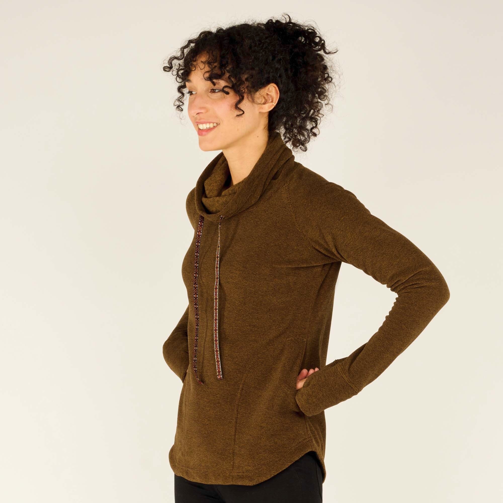 A close-up side profile of the woman, showing a detailed view of the cowl neck with the drawstring ties and the soft texture of the Sherpa Adventure Gear Rolpa Pullover in Light Brown fabric. She is smiling and has her hands in the pockets of the pullover, emphasising its relaxed and comfortable fit.