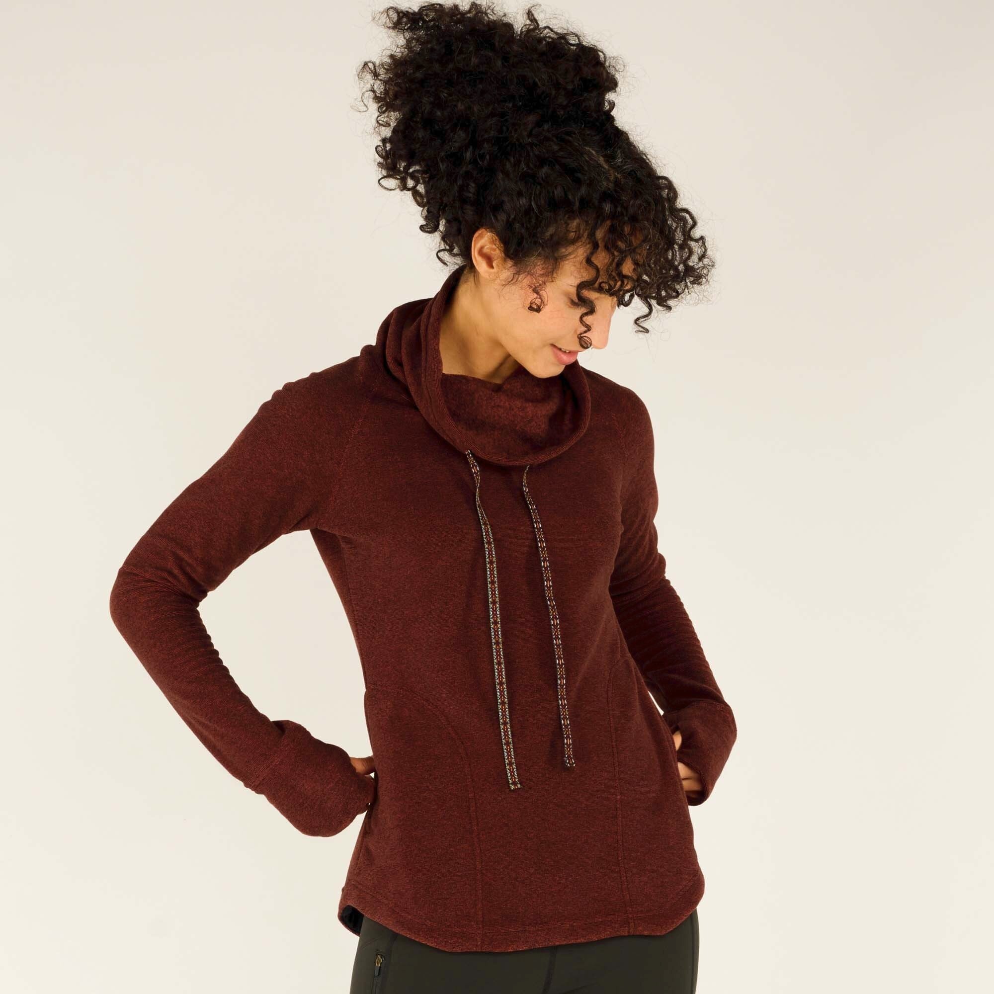 A female model with curly dark hair wears the Sherpa Adventure Gear Rolpa Pullover in Red. She is shown from the front, posing with one hand on her hip and the other tucked into her front kangaroo pocket. The pullover features a cozy cowl neck with two decorative drawstrings, and the fabric has a soft heathered texture. She pairs the pullover with dark green leggings.