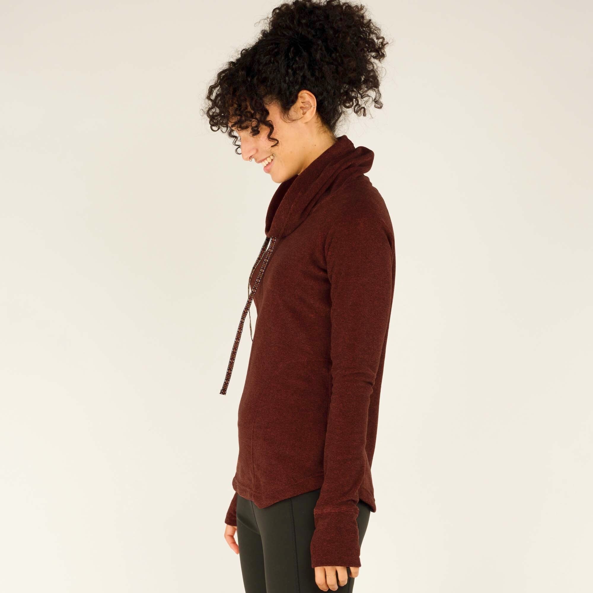 A side profile of the model highlights the Sherpa Adventure Gear Rolpa Pullover in Red’s relaxed fit and the way the cowl neck gently drapes down the front. The curved hem is slightly longer in the back, providing extra coverage. The sleeves feature thumbholes for added coziness, and the fabric’s soft texture is visible.