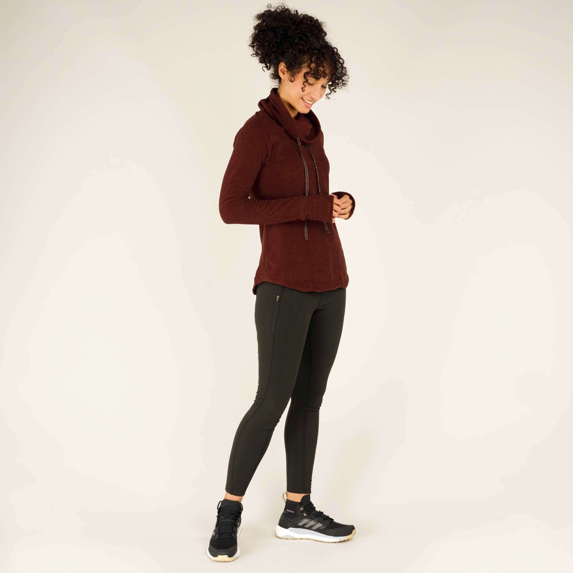 A three-quarter angle shot of the model captures her smiling softly while looking down at her hands. The Sherpa Adventure Gear Rolpa Pullover in Red’s drawstrings, front pocket, and thumbholes are all visible, highlighting the pullover’s practical and stylish design. The combination of the deep red pullover with the dark green leggings creates a harmonious, nature-inspired color palette.