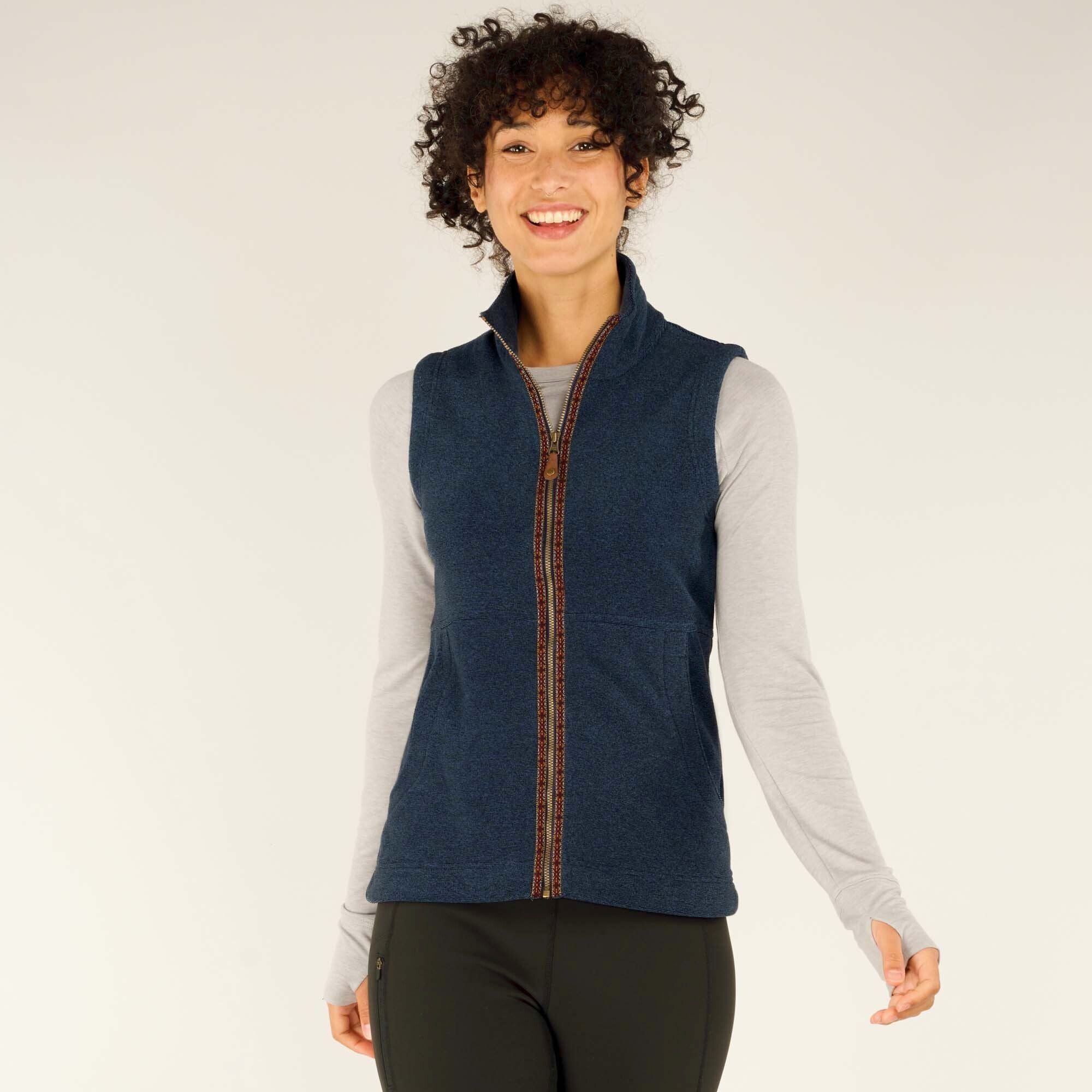 A smiling woman with curly hair models a Sherpa Adventure Gear Rolpa Vest in Blue over a light gray long-sleeve shirt. The vest features a high collar, a full-length zipper with intricate woven trim, and two front pockets. She is posing against a neutral background, with her right arm slightly extended and her left hand relaxed at her side.