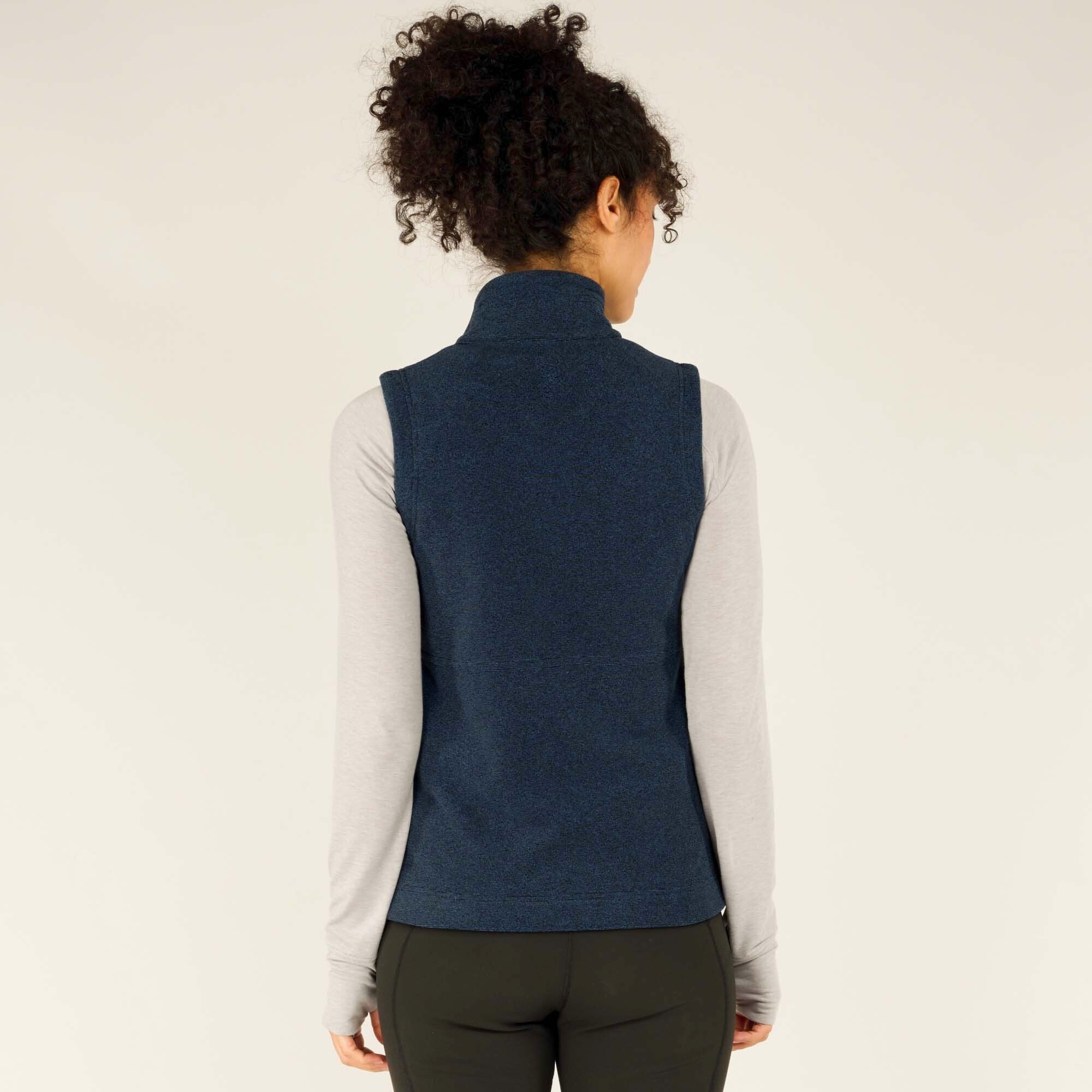 A back view of the model wearing the Sherpa Adventure Gear Rolpa Vest in Blue. The vest’s simple design is visible, with a high collar and a smooth back panel. The model’s curly hair is styled naturally, and she stands with a relaxed posture against a plain background.