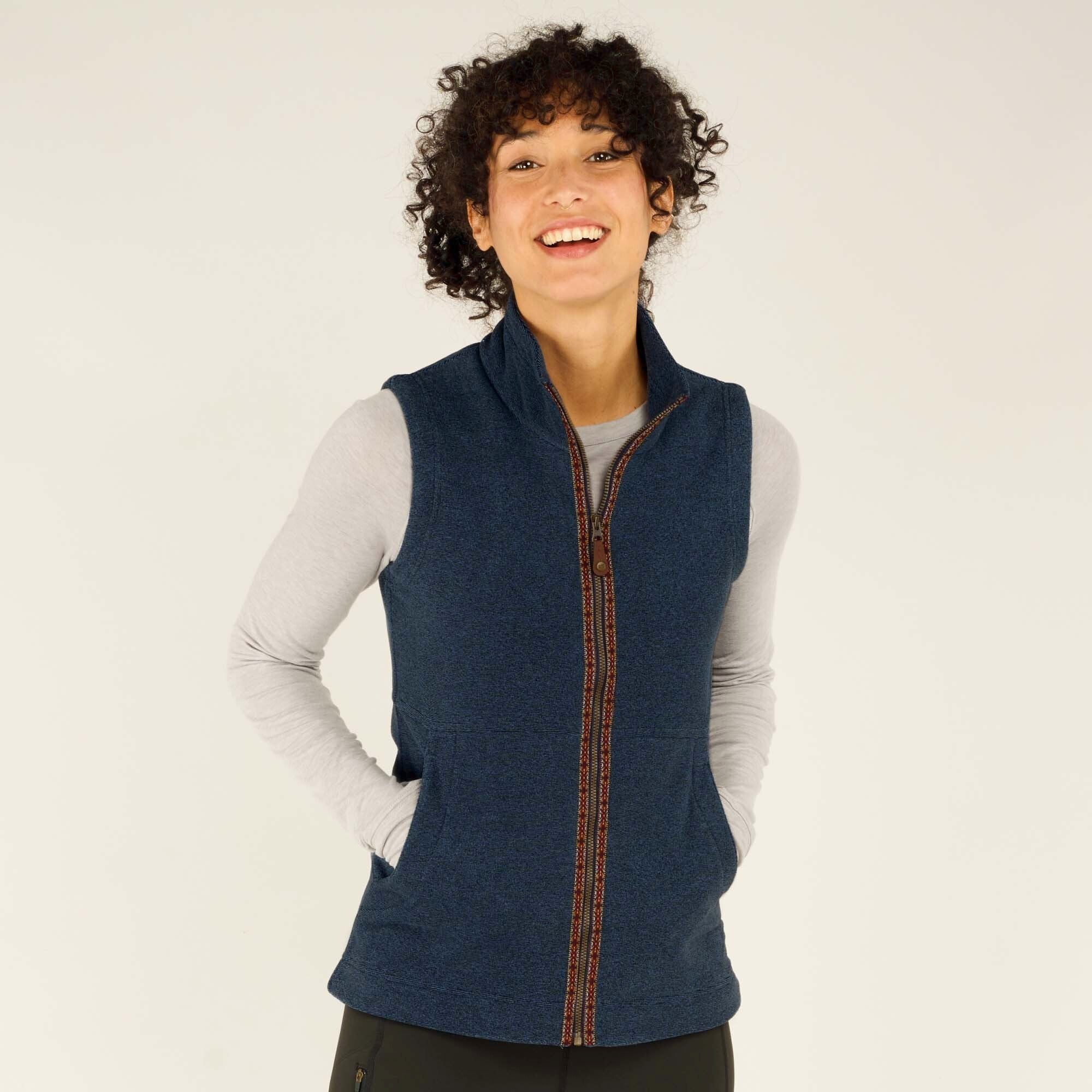 A close-up of the model smiling while wearing the Sherpa Adventure Gear Rolpa Vest in Blue. She places both hands in the vest’s front pockets, showing the warmth and comfort of the garment. The woven trim along the zipper is visible, and she looks cheerful against the neutral backdrop.