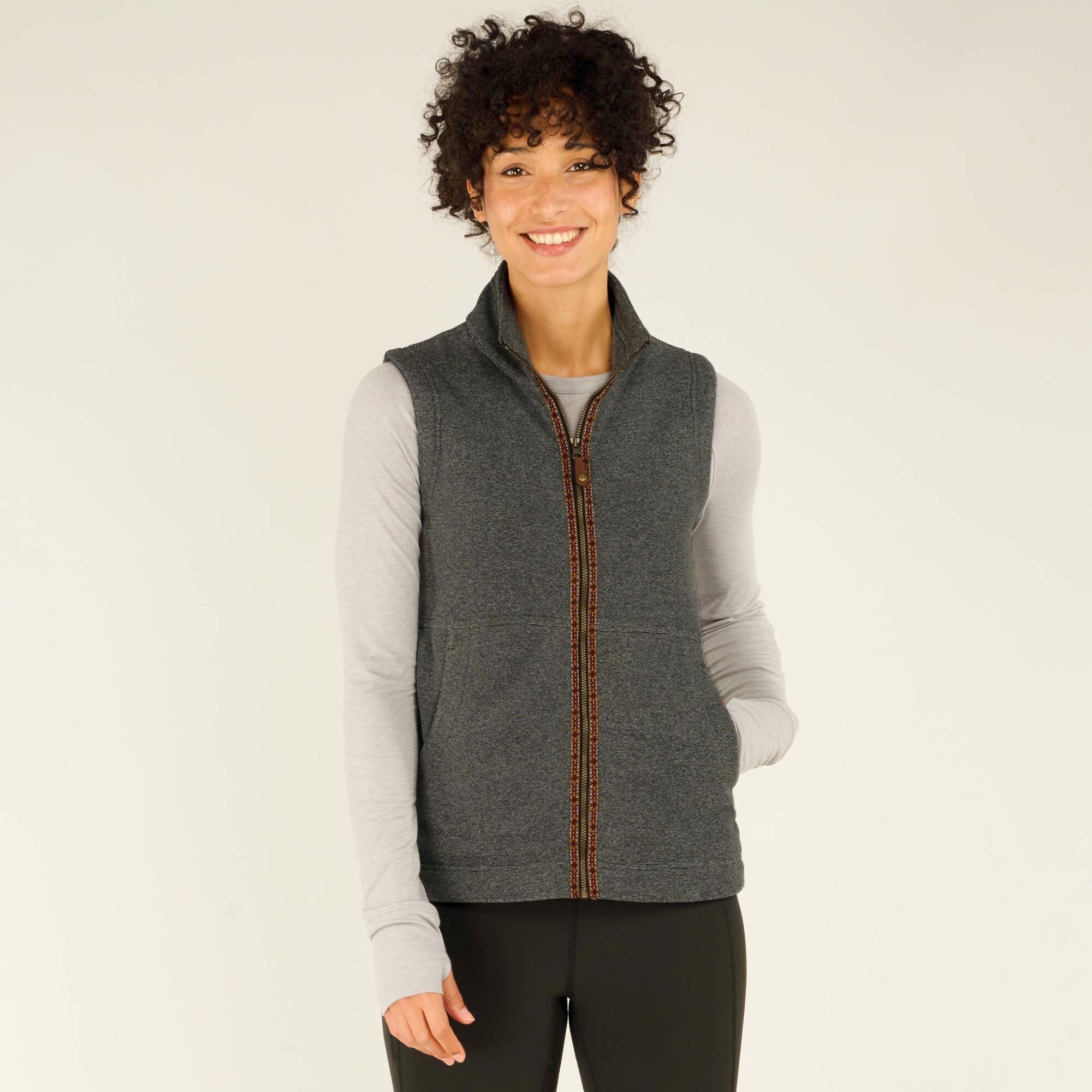 A smiling woman with short, curly dark hair is wearing a Sherpa Adventure Gear Rolpa Vest in Grey over a light grey long-sleeve shirt. The vest has a high collar and two front pockets. She stands against a neutral beige background with her hands in the pockets, looking directly at the camera.