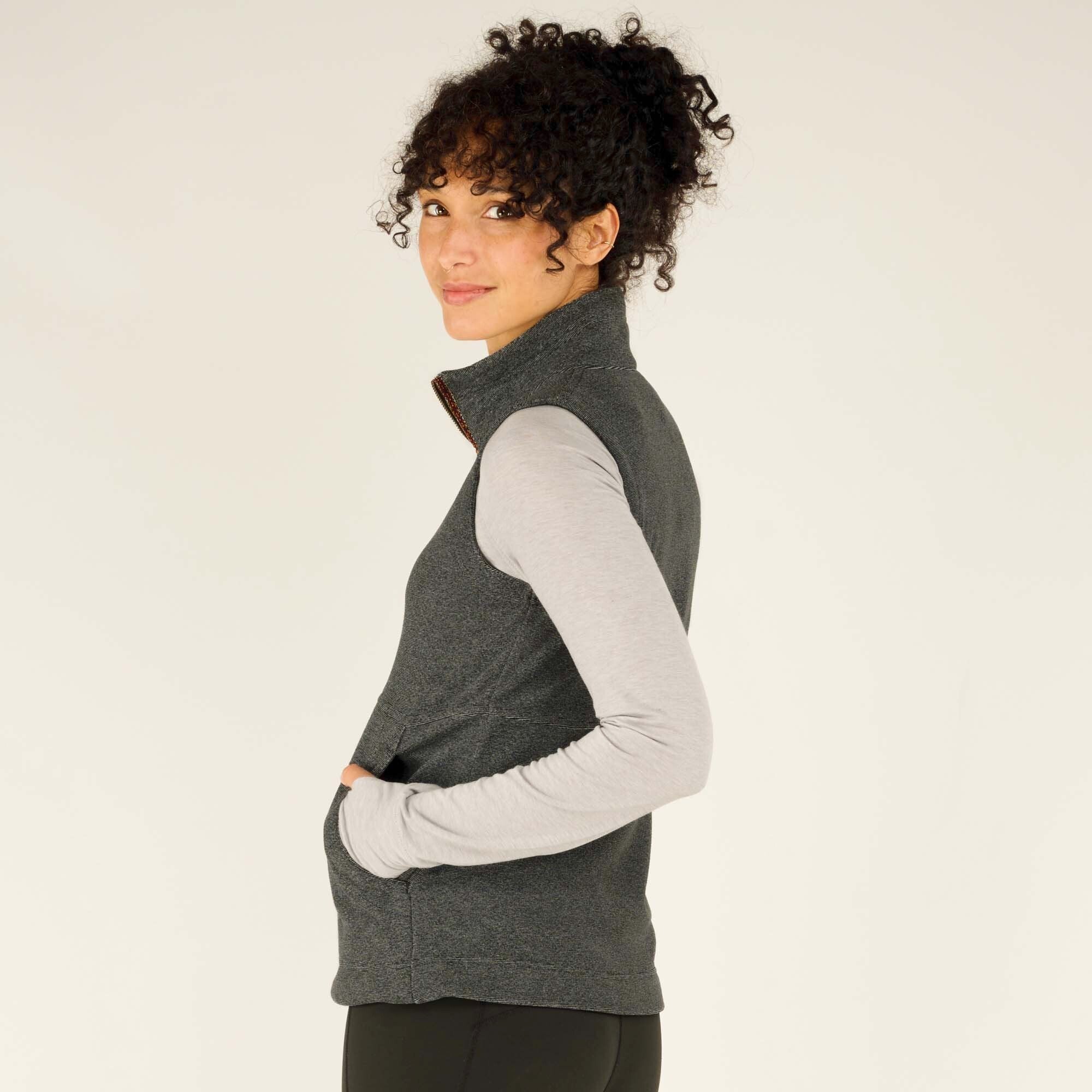 A profile view of the woman in the Sherpa Adventure Gear Rolpa Vest in Grey, showing the fit of the vest and its raised collar. She places her hands in the vest pockets while gazing slightly back at the camera with a subtle smile.