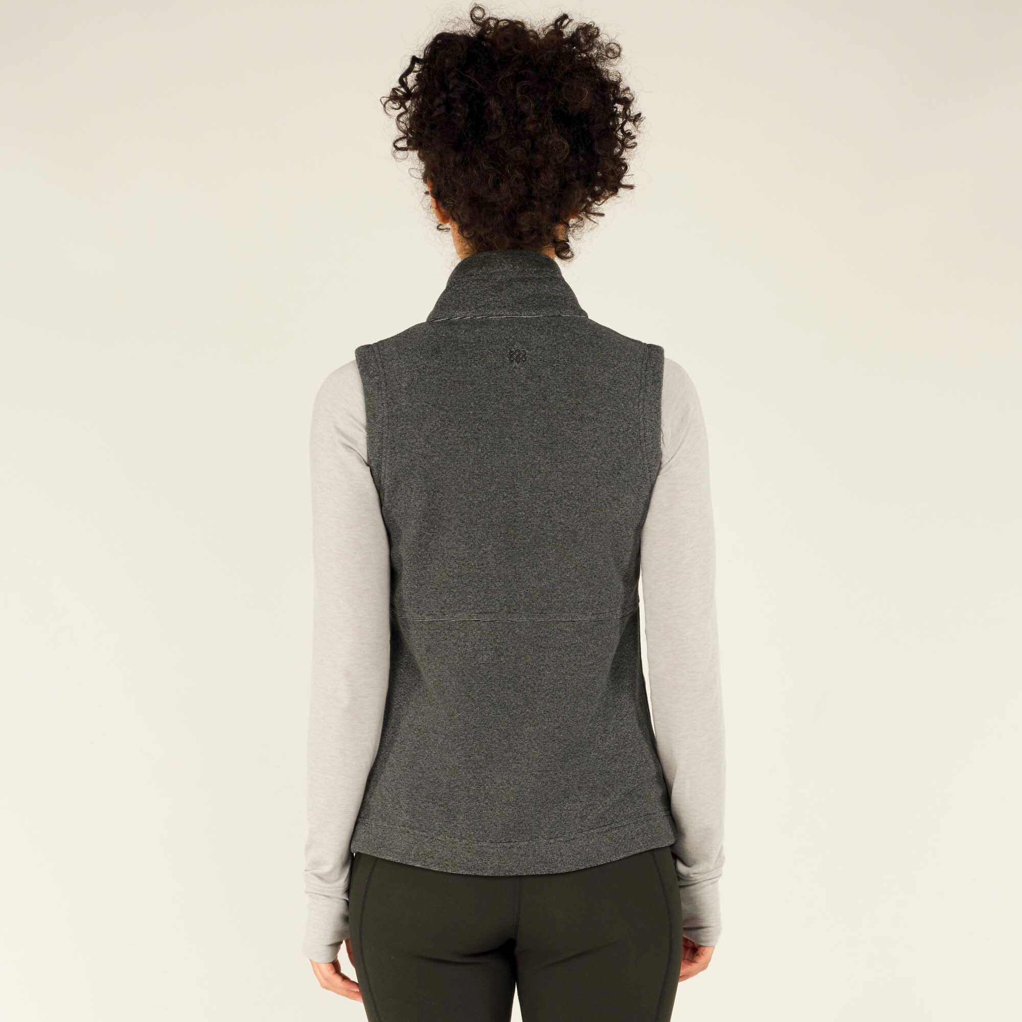 The back view of the Sherpa Adventure Gear Rolpa Vest in Grey, highlighting its simple design, high collar, and fitted cut. The woman stands with her arms at her sides, showcasing the seamless structure and length of the vest.