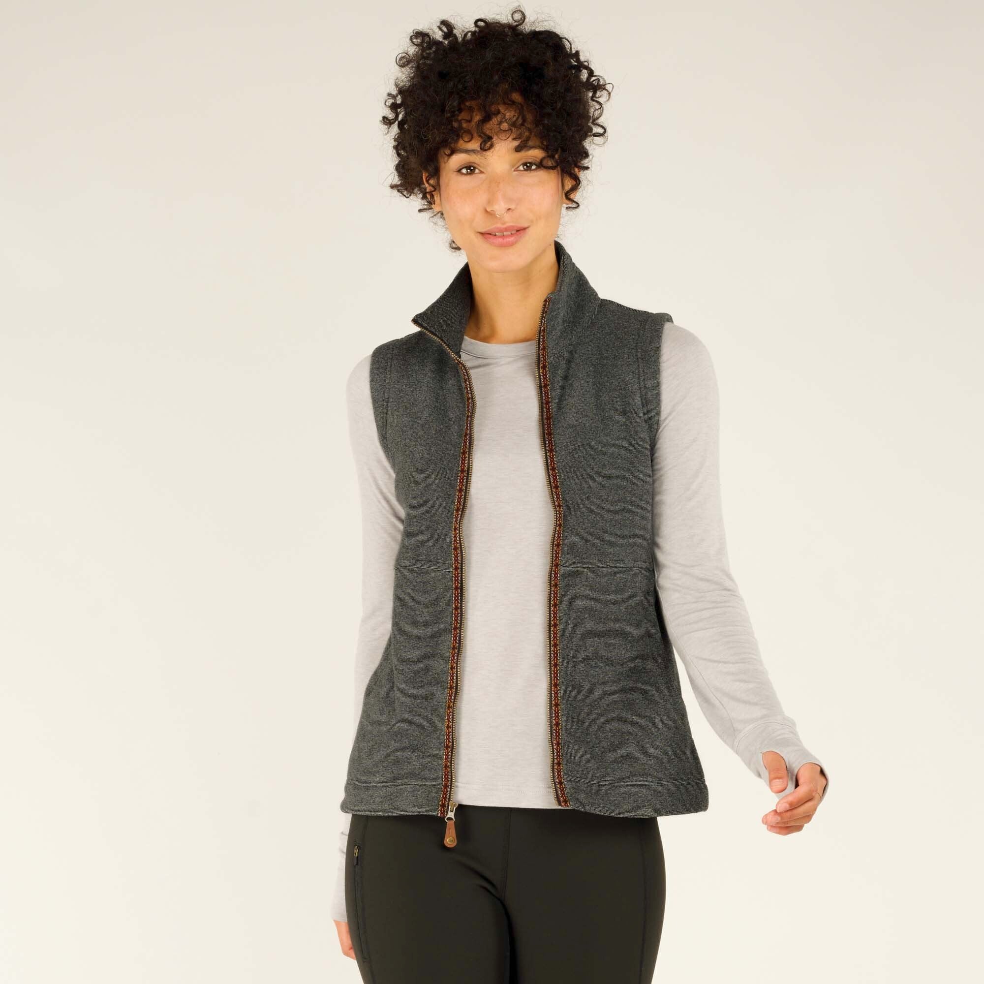 The woman is wearing the same Sherpa Adventure Gear Rolpa Vest in Grey, now unzipped to reveal the full-length zipper with decorative stitching along the edges. She holds the edges of the vest slightly apart, standing in a relaxed stance while smiling.