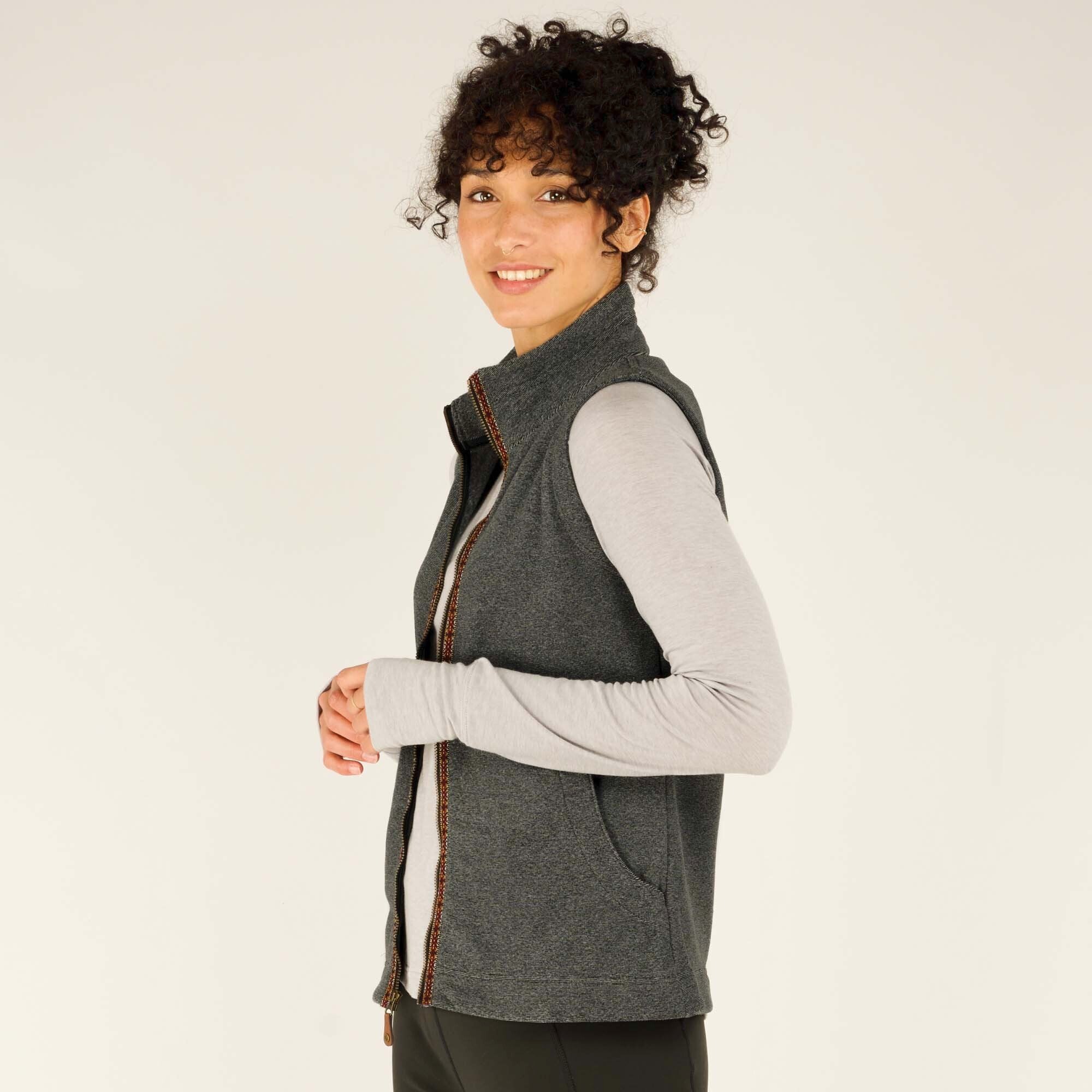 A side angle of the unzipped Sherpa Adventure Gear Rolpa Vest in Grey, revealing the interior lining. The woman, still in her long-sleeve light grey shirt, smiles warmly while turning slightly toward the camera, showing the vest's side pockets and smooth texture.