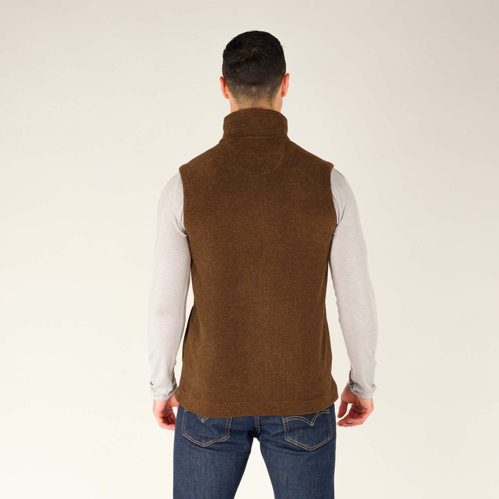 A rear view of the model wearing the Sherpa Adventure Gear Rolpa Vest in Light Brown, showcasing the high collar and the vest’s smooth, seamless back design. The vest is paired with dark blue jeans, and the background remains neutral.