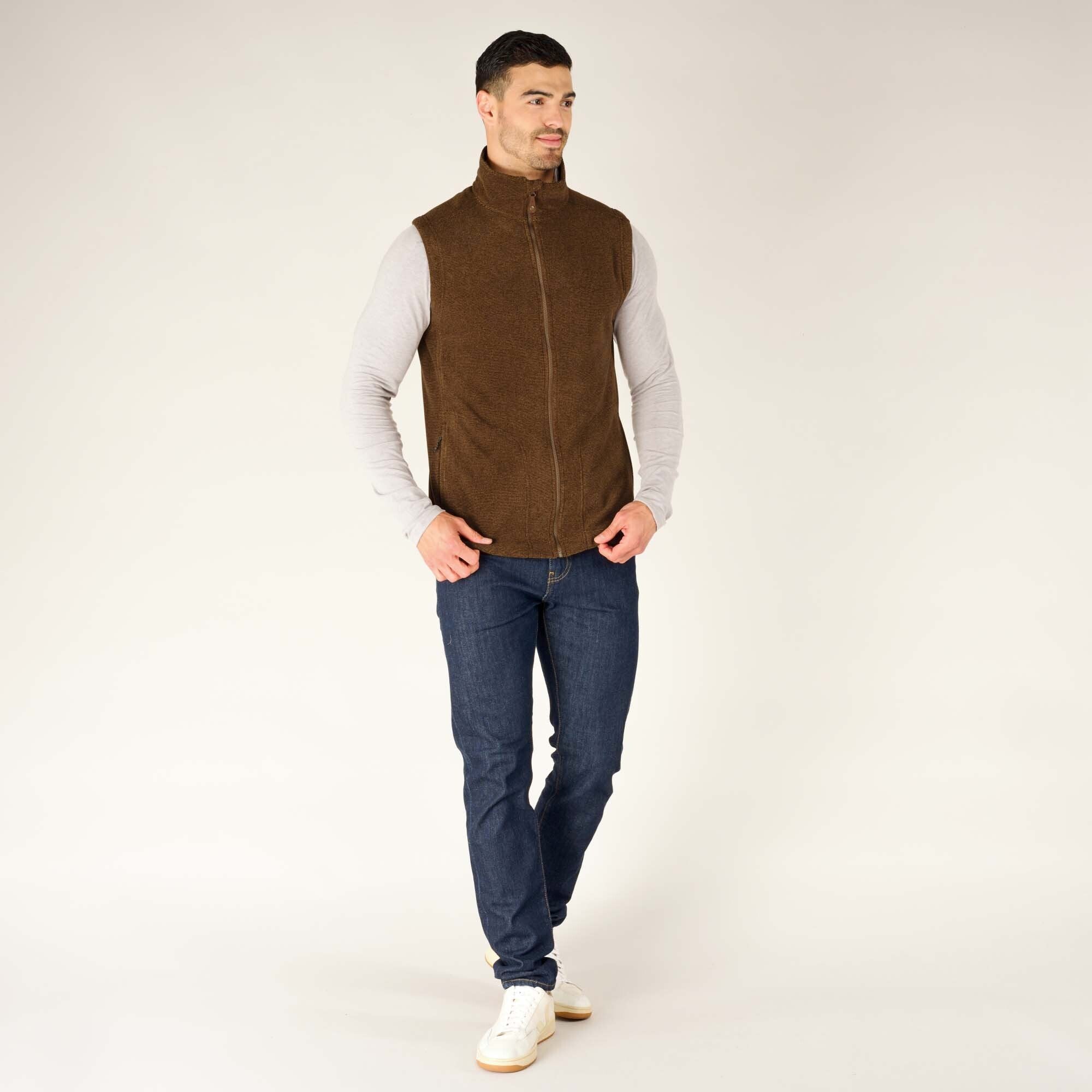 The model wearing the Sherpa Adventure Gear Rolpa Vest in Light Brown, styled with dark blue jeans and white trainers. He is standing with a slight lean and hands in the vest pockets, smiling while facing slightly to the right. The neutral background highlights the vest’s earthy tone.