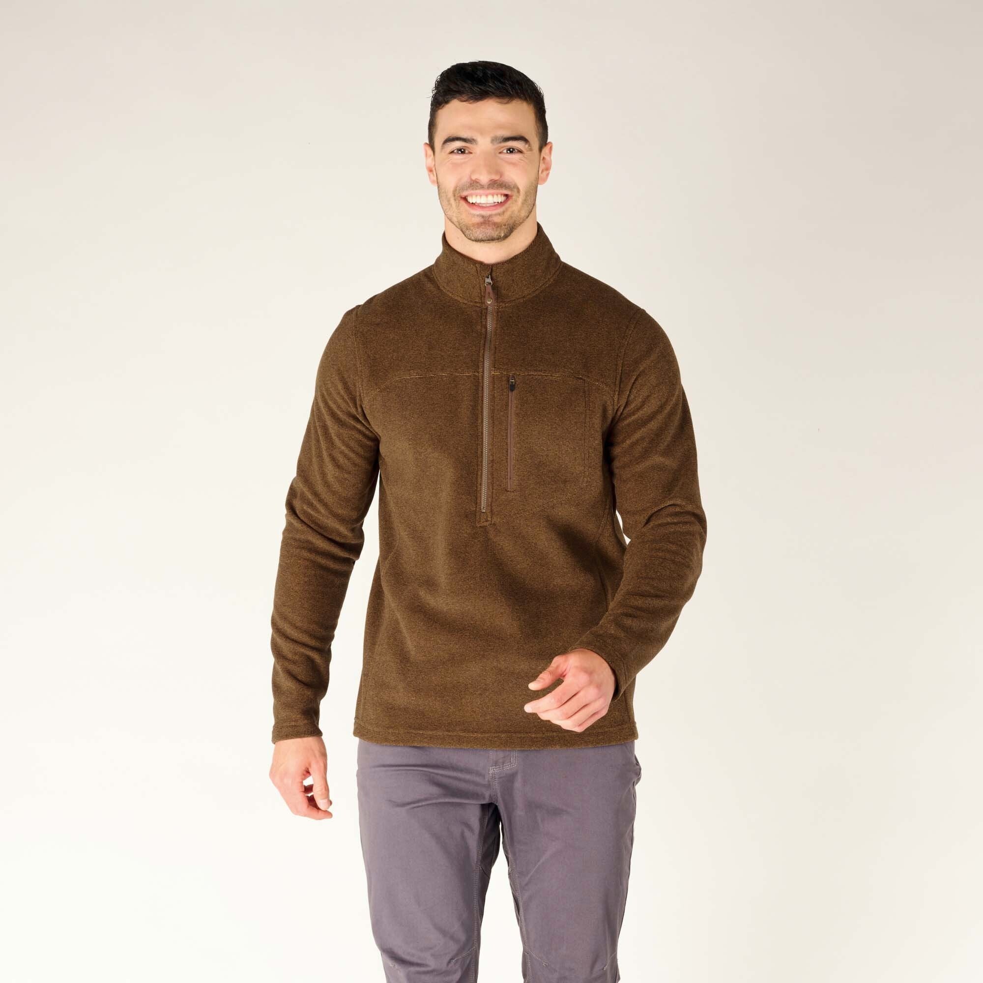 A male model with short dark hair and a beard is wearing a Sherpa Adventure Gear Rolpa Zip T-Shirt in Light Brown with a half-zip front closure and a chest pocket with a zipper. He is smiling at the camera with a relaxed pose, one hand slightly extended. He is also wearing gray pants, and the background is a plain, off-white color.