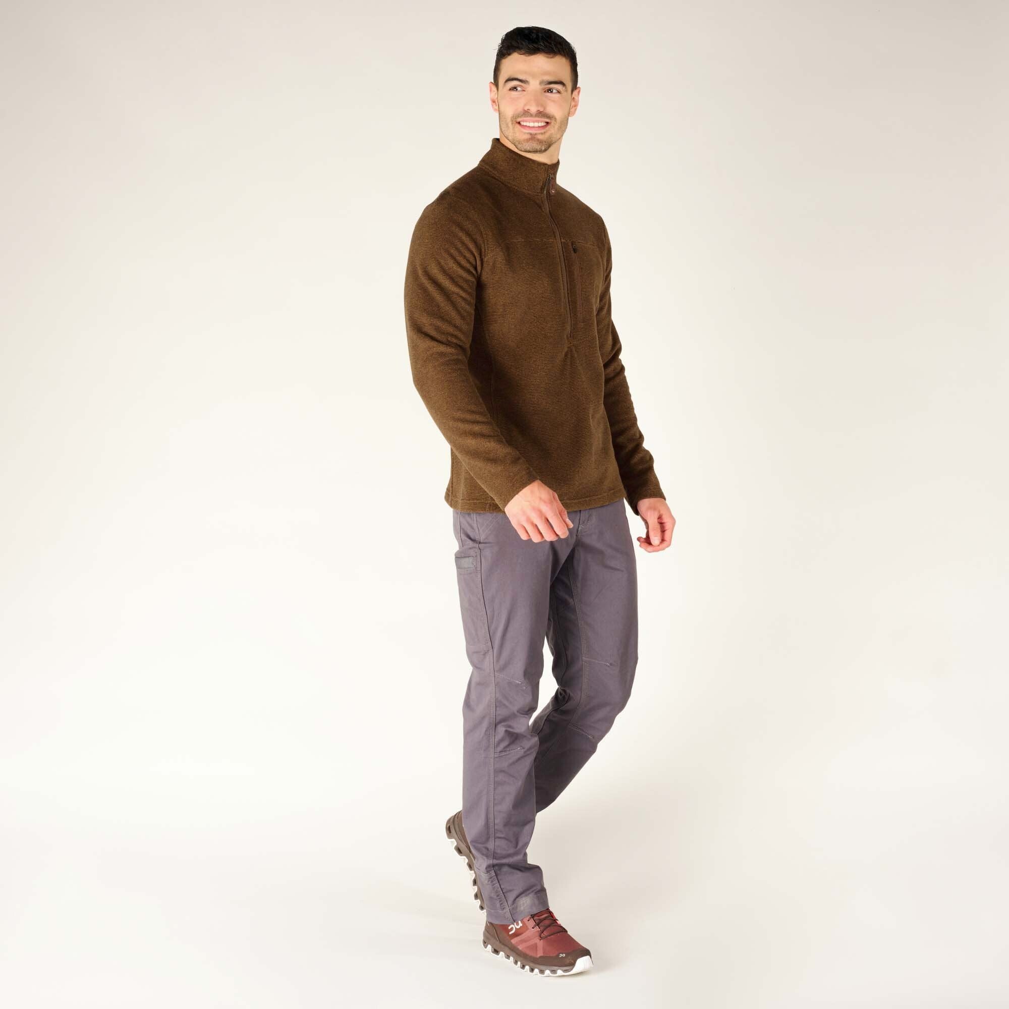 The male model is captured mid-step, wearing the Sherpa Adventure Gear Rolpa Zip T-Shirt in Light Brown and gray pants. His head is slightly turned, showing a smile, and his left hand is tucked into his pocket. The side profile highlights the relaxed fit and length of the shirt, while he is also wearing brown hiking shoes.