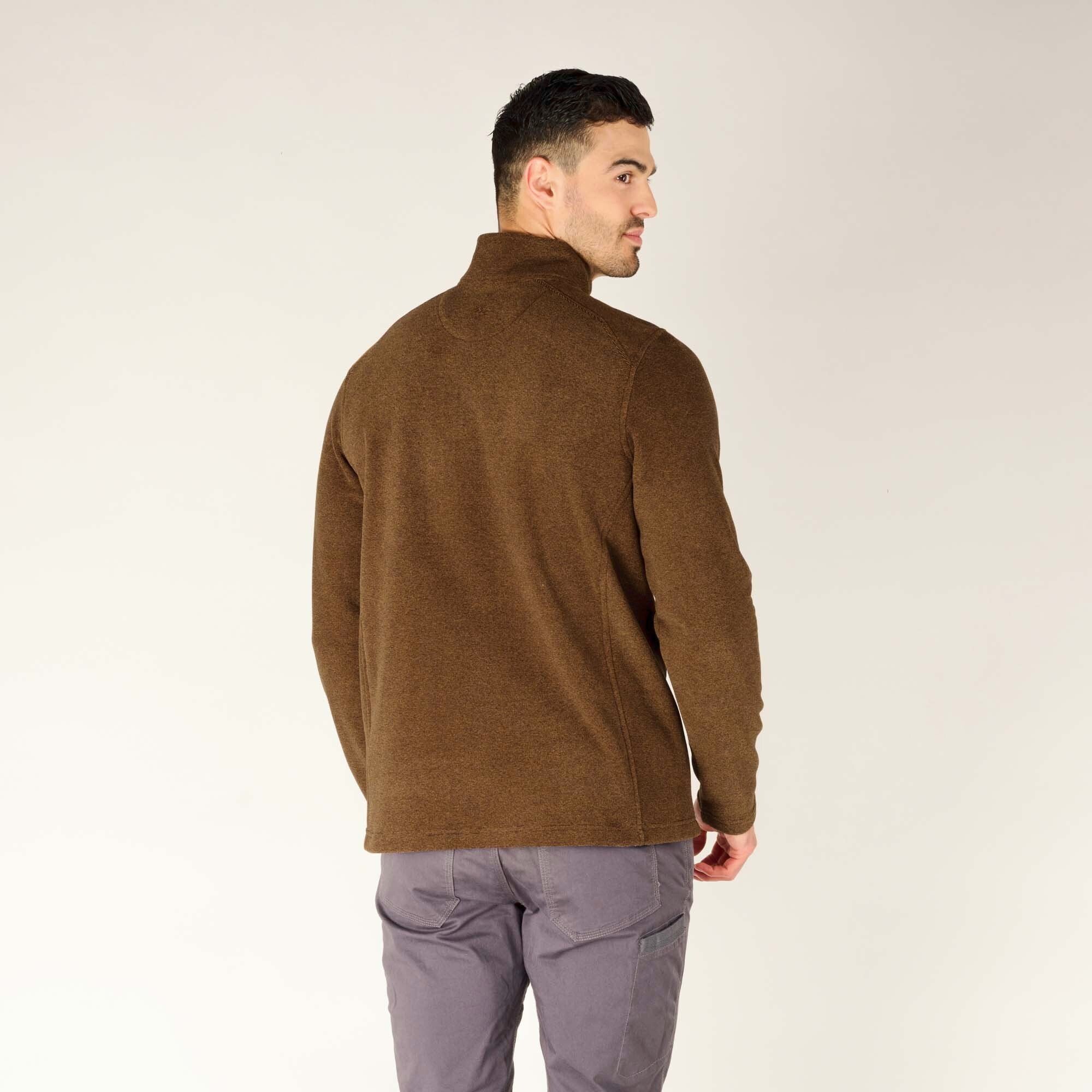 A rear view of the model wearing the Sherpa Adventure Gear Rolpa Zip T-Shirt in Light Brown, showing the simple design of the back with no visible branding. The high collar provides coverage, and the hem falls just below the waist. The gray pants and brown hiking shoes complete the outfit.