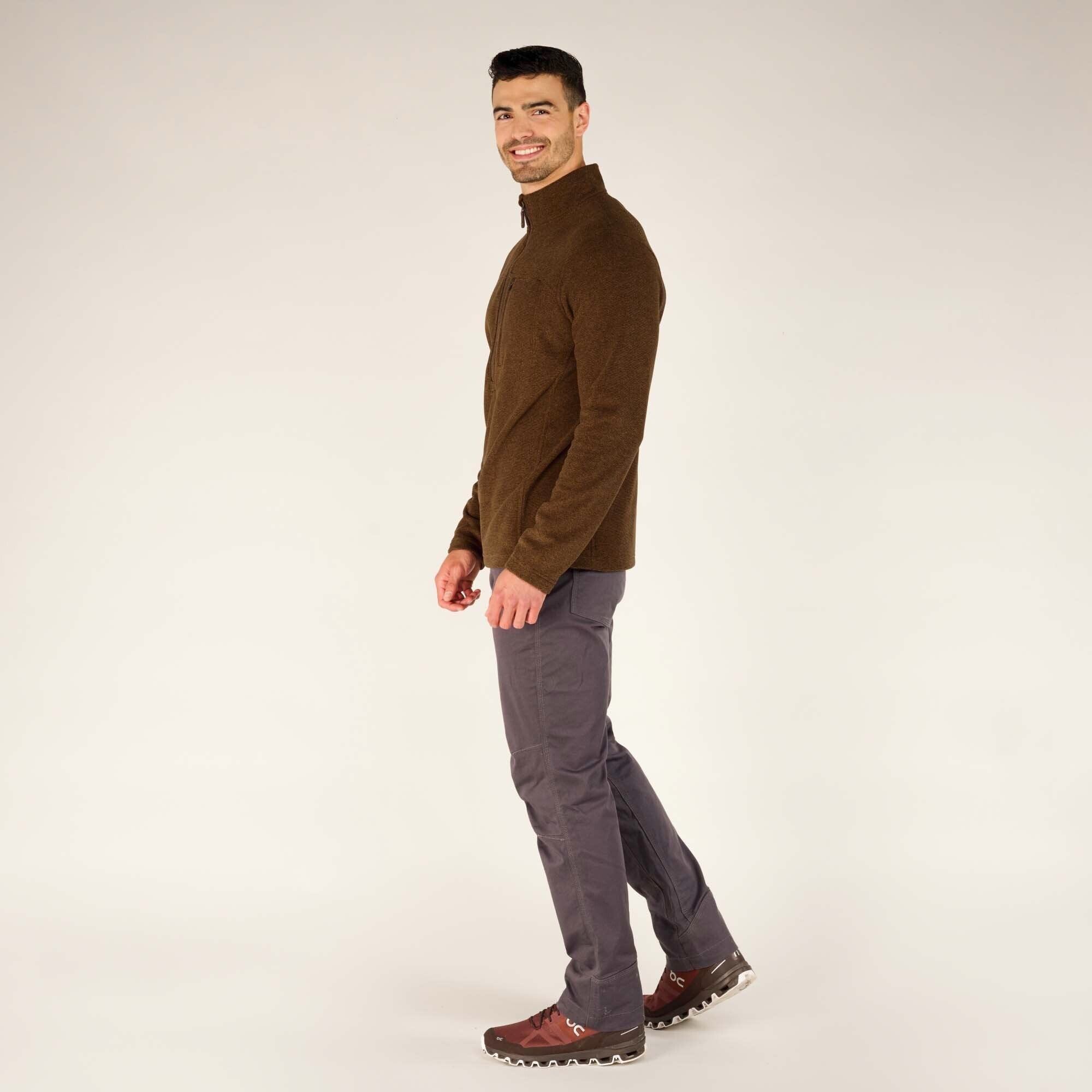 The male model is shown from the side, wearing the Sherpa Adventure Gear Rolpa Zip T-Shirt in Light Brown, gray pants, and brown hiking shoes. He is smiling and looking toward the camera. The side view emphasizes the fit of the tee, which sits comfortably over the pants.