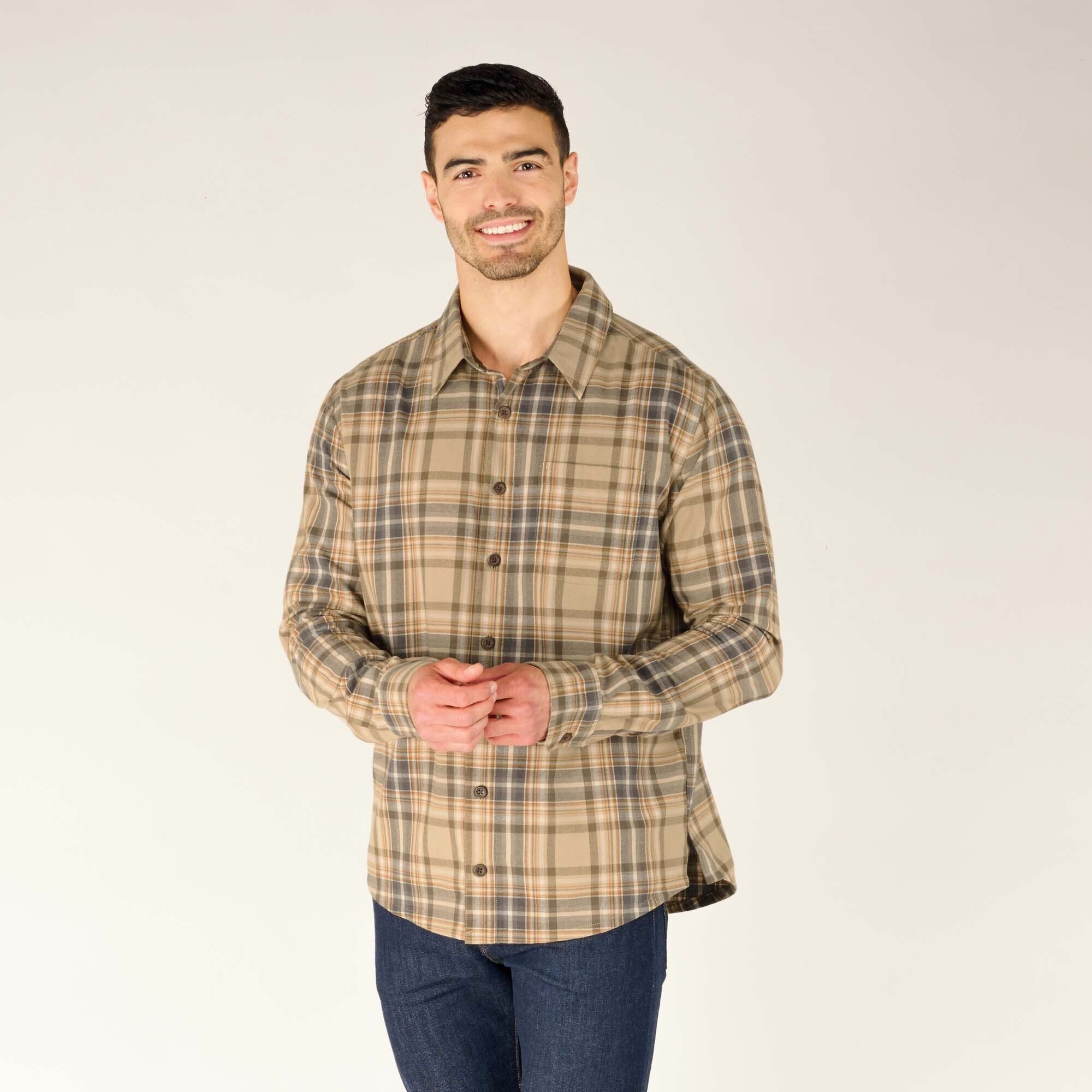 A male model with short dark hair and a well-groomed beard is wearing a Sherpa Adventure Gear Rudra Shirt in Beige. He stands with a relaxed posture, smiling, and lightly clasping his hands in front of him. He is also wearing dark blue jeans. The background is a neutral beige.