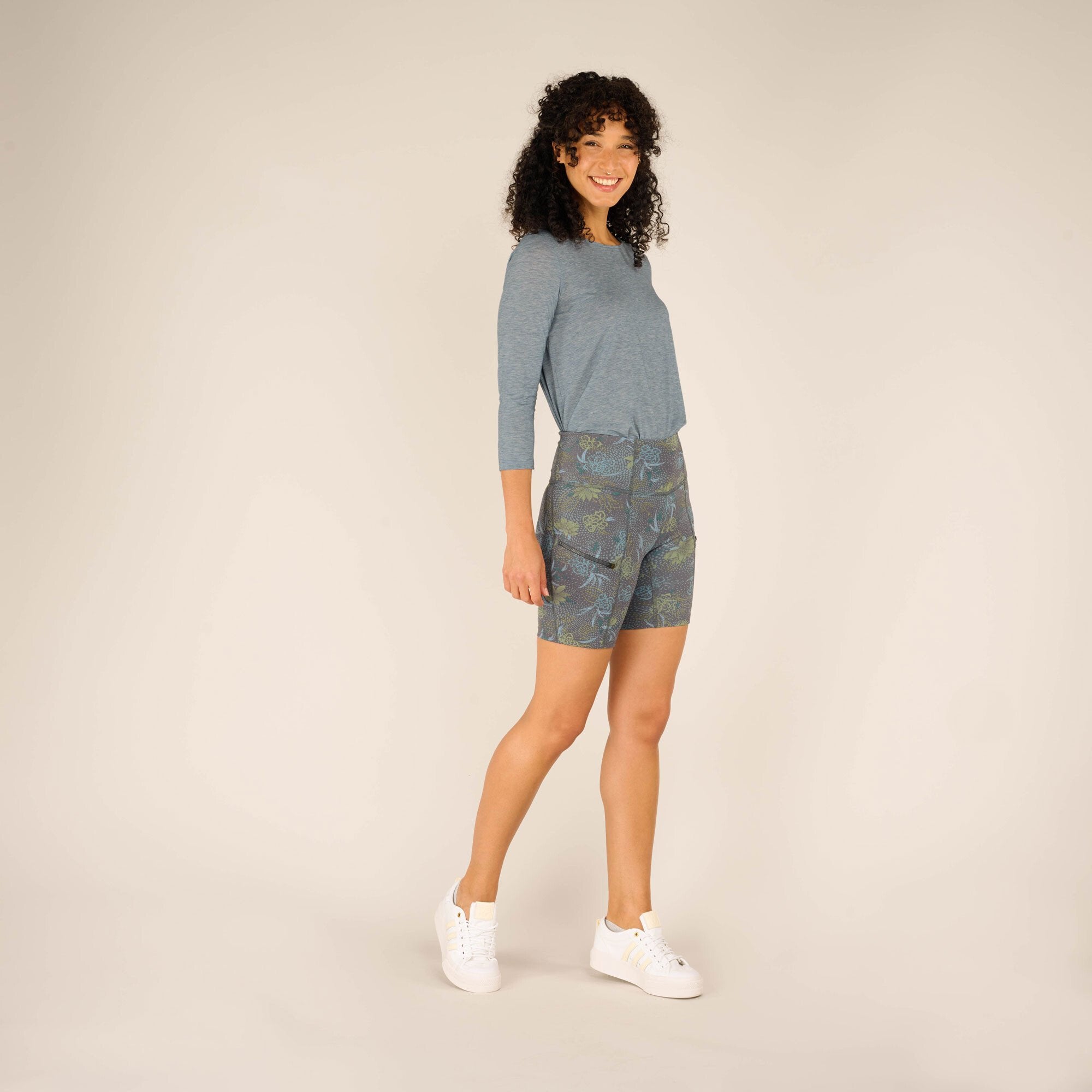 The model stands angled to the left, showcasing the contouring fit of the Sherpa Adventure Gear Sahasa Shorts in Blue and their floral pattern. The outfit highlights the streamlined silhouette with a tucked-in light blue top, exuding a sporty and comfortable style.