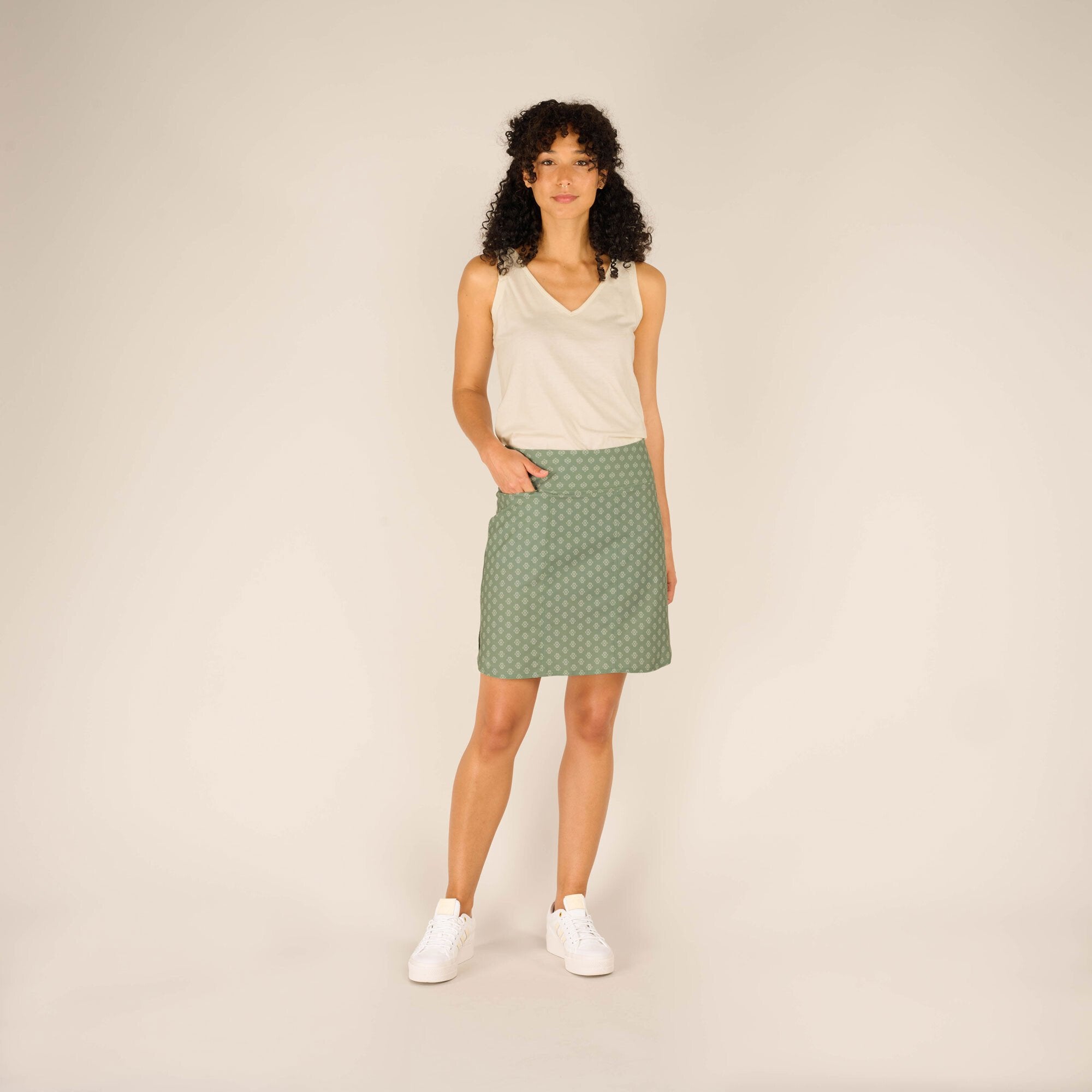 The model is standing in a relaxed pose, showing the Sherpa Adventure Gear Sajilo Adventure Skort in Green paired with a beige sleeveless top and white trainers, creating a casual yet stylish look.