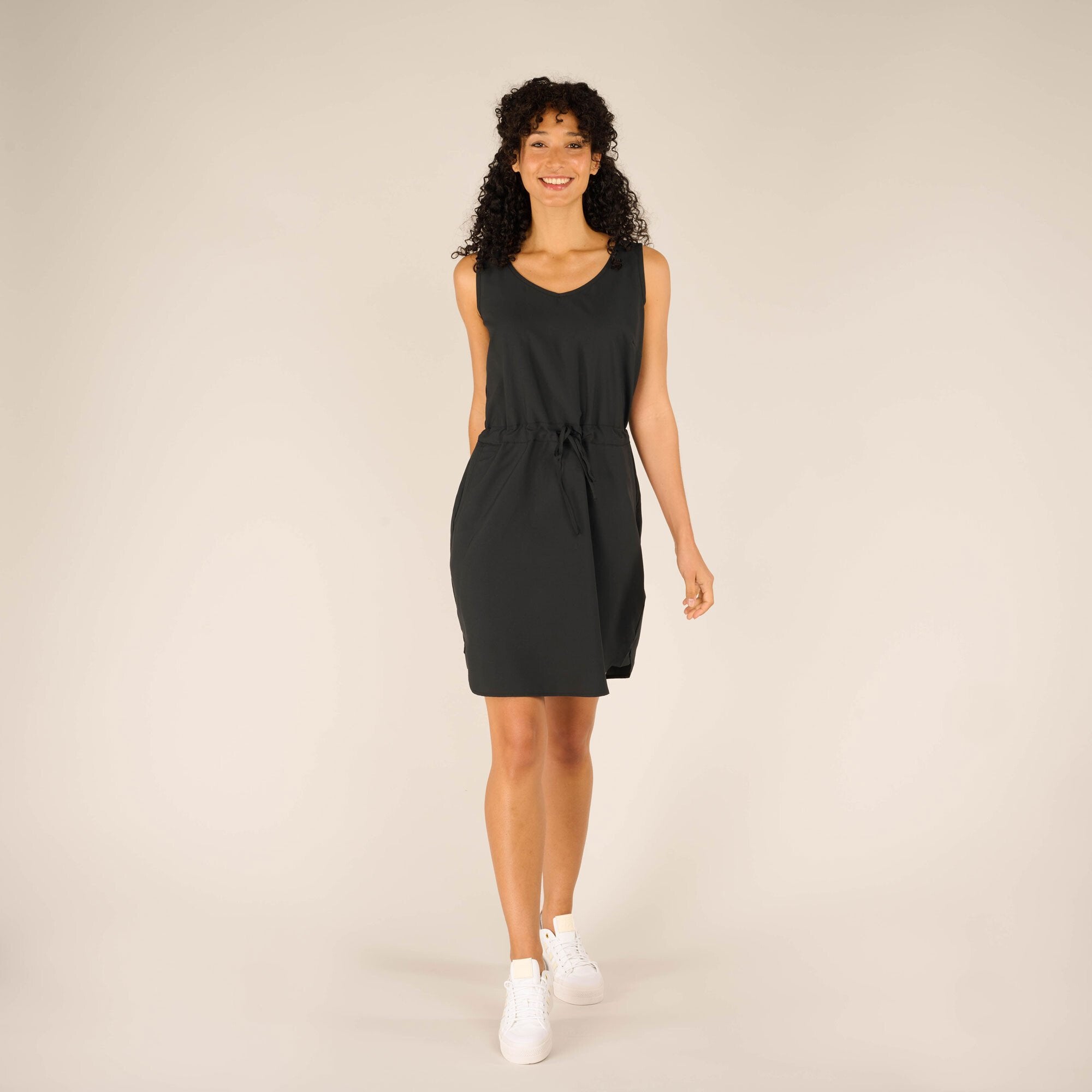 A front view of a model wearing a Sherpa Adventure Gear Sajilo Dress in Black with a relaxed fit and a cinched drawstring waist. The dress has a V-neckline and two side pockets. The model is walking forward with a cheerful expression, paired with white trainers.