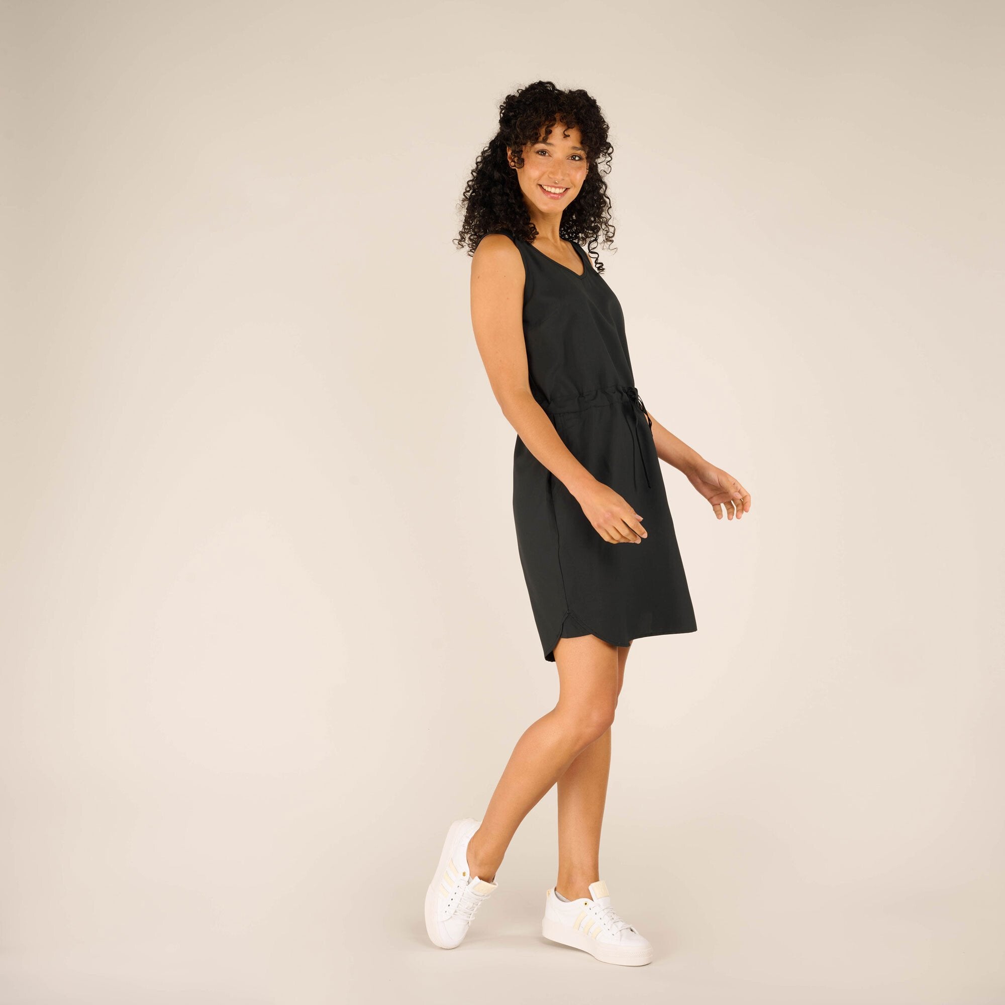 A side view of the Sherpa Adventure Gear Sajilo Dress in Black showing the flowy fabric and casual silhouette. The model is turned slightly to her left, smiling while looking over her shoulder, showcasing the dress's versatility for casual settings.