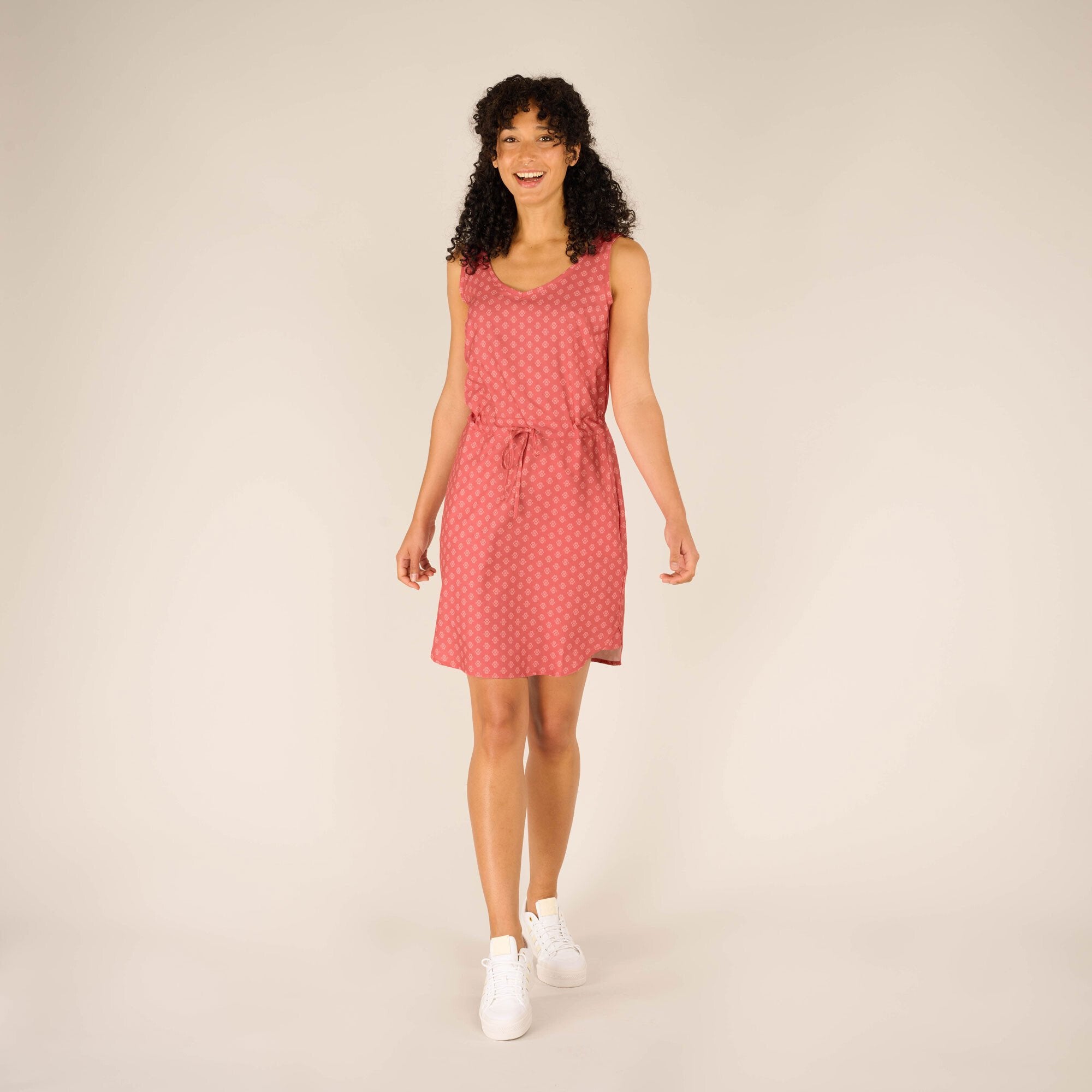 A woman wearing a Sherpa Adventure Gear Sajilo Dress in Pink with a subtle geometric pattern. The dress has a V-neckline, a waist tie for adjustable fit, and ends just above the knee. She is standing in a neutral pose with white trainers, smiling brightly, and her curly hair frames her face beautifully.