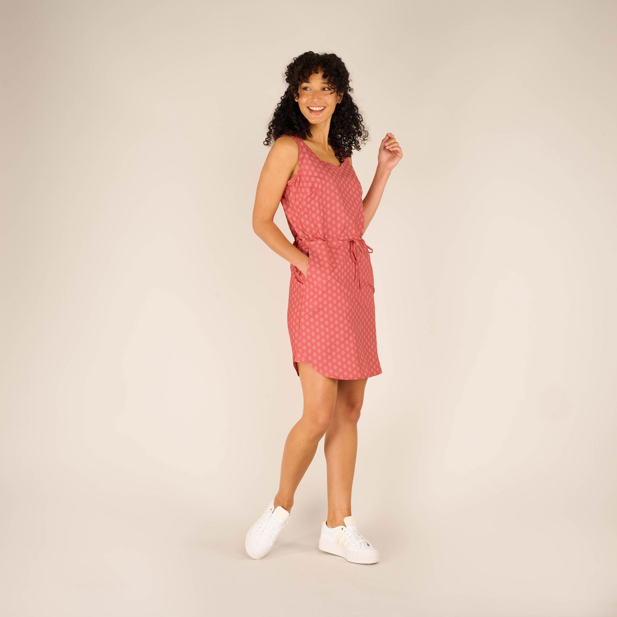 The same woman poses with one hand placed casually in her Sherpa Adventure Gear Sajilo Dress in Pink pocket. The waist tie detail is prominent, and the side slits on the hem of the dress add a stylish touch. Her white trainers complement the casual look, and she maintains a confident smile.