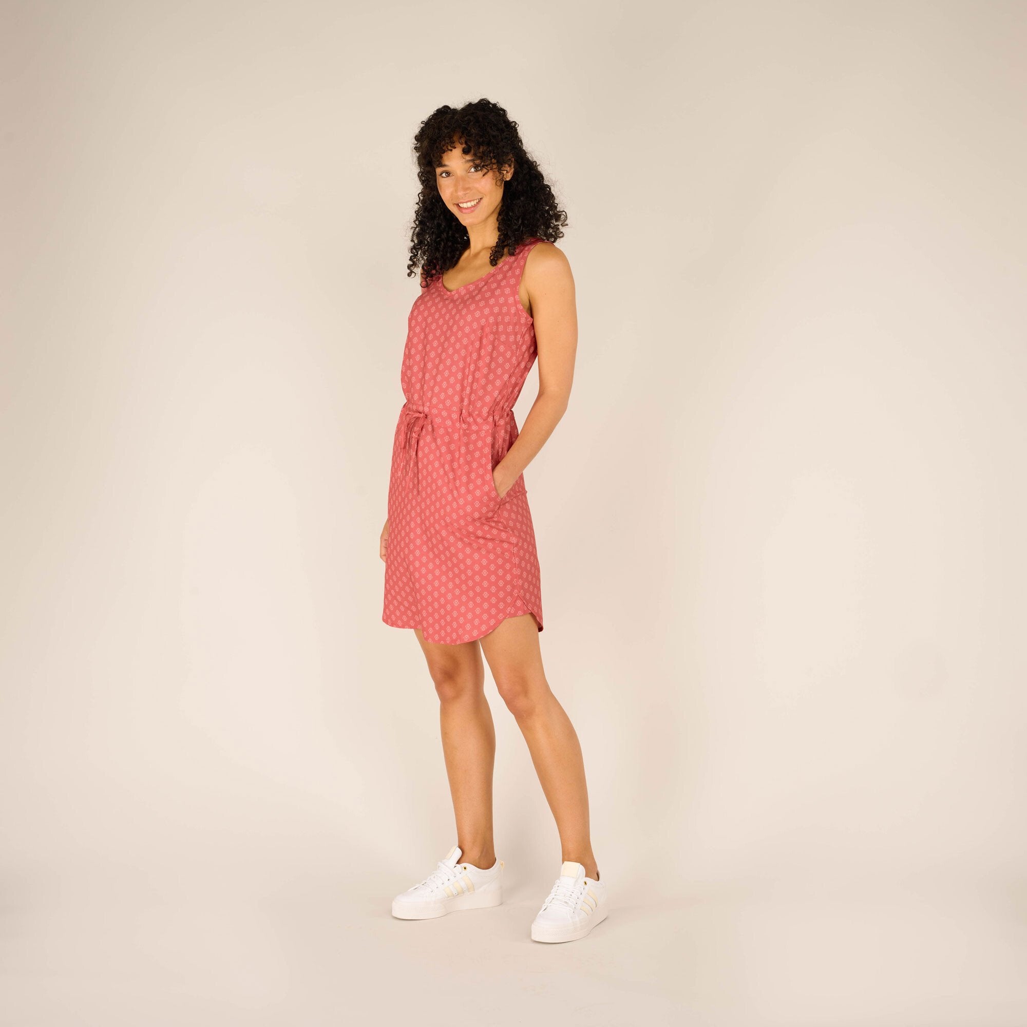 A full-body front view of the woman, standing confidently with a slight tilt to one side. The overall fit of the Sherpa Adventure Gear Sajilo Dress in Pink and its casual, comfortable vibe are emphasised. The white trainers add a sporty and relaxed touch.