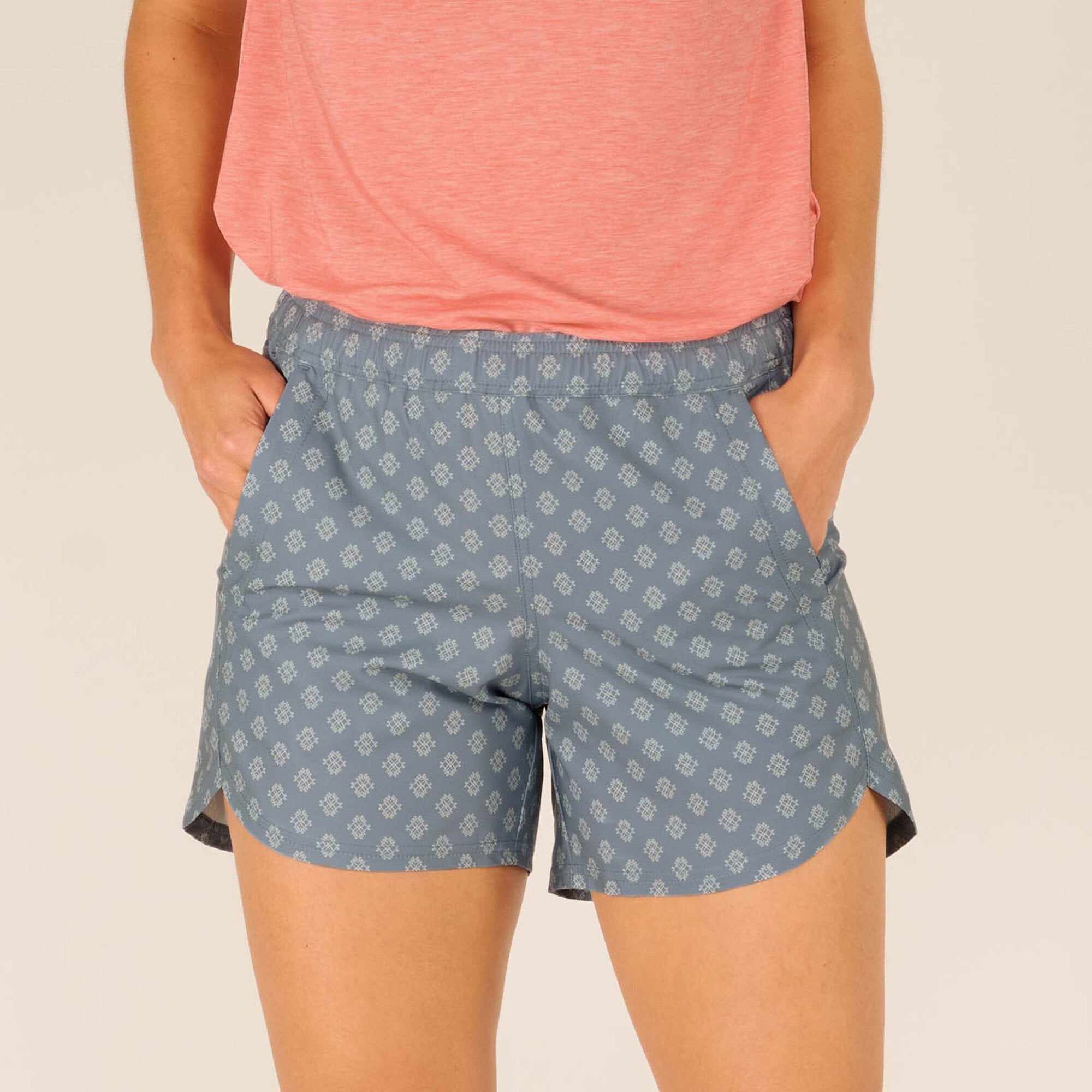 The image shows a close-up of the Sherpa Adventure Gear Sajilo Pull-On Shorts in Blue with a subtle geometric pattern. The shorts feature a comfortable elastic waistband, two front pockets, and small side slits for ease of movement. The model is wearing a coral pink top, partially tucked in, showcasing the shorts' fit and waistband.