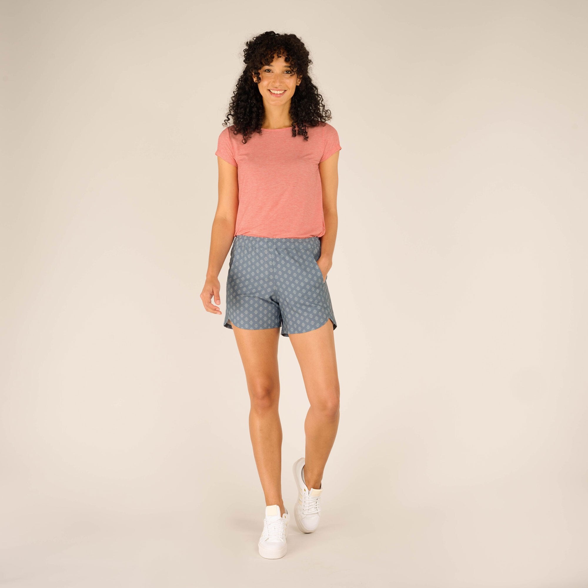 The model is wearing the Sherpa Adventure Gear Sajilo Pull-On Shorts in Blue paired with a coral pink short-sleeve top and white trainers. This full-body image highlights the casual and comfortable fit of the shorts, including their slightly relaxed style and functional pockets.